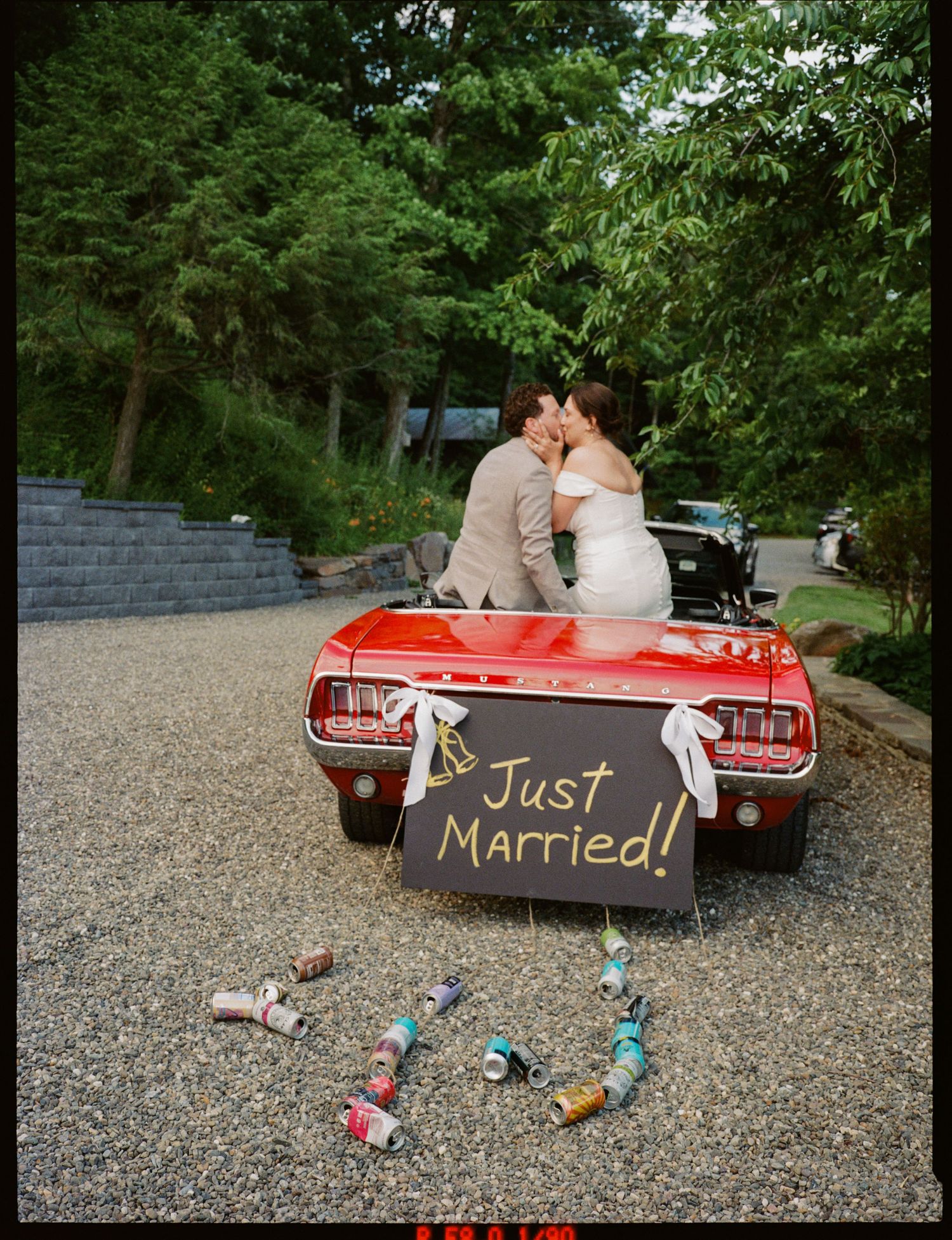 Newlyweds kissing on a red convertible with a Just Married sign and tin cans trailing behind.