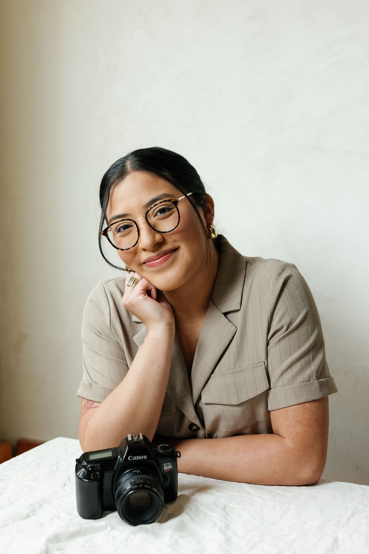 Professional photographer wearing beige blazer poses with camera on white bedding in minimalist portrait series.