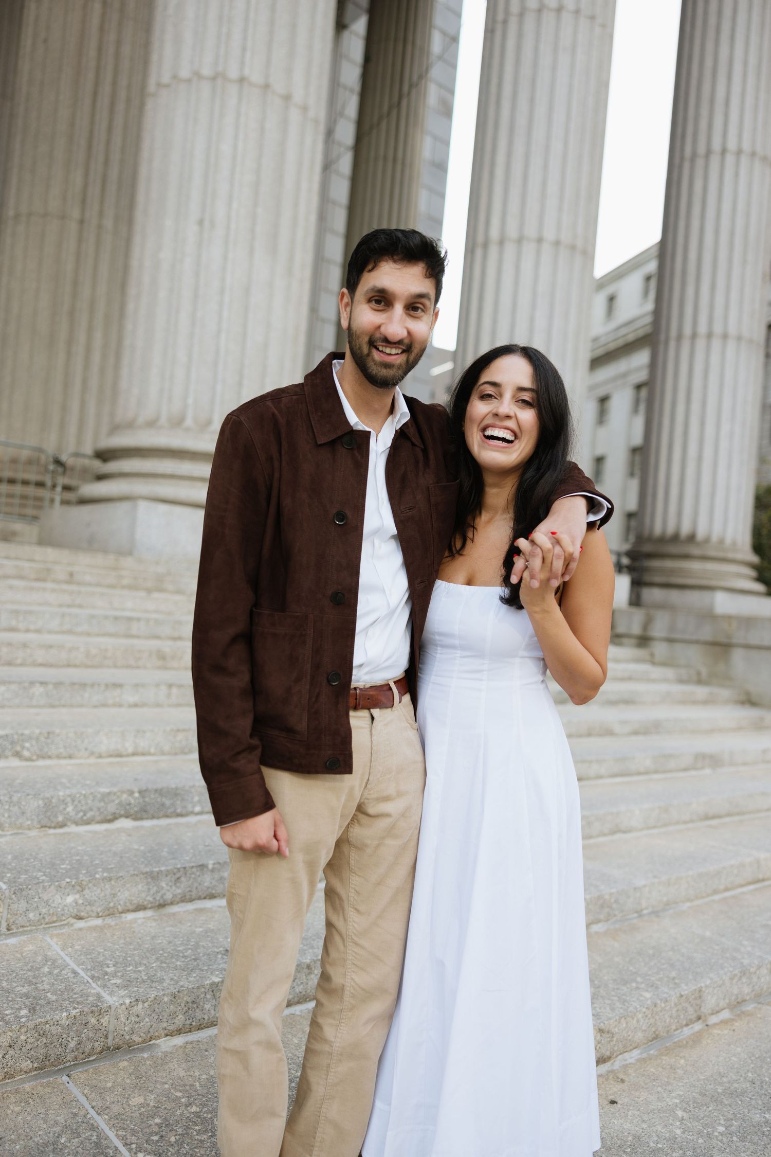 A couple shares joyful laughter together while posing in front of classic white stone columns.