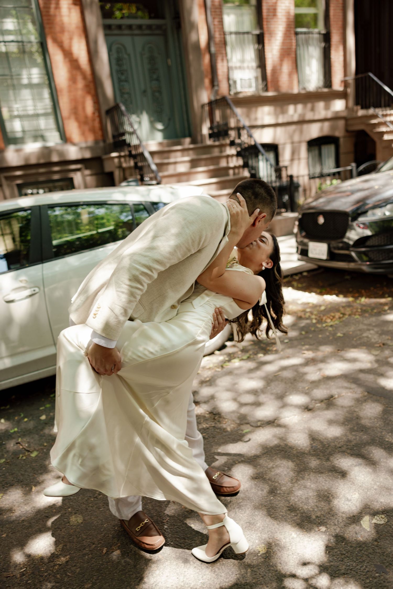 Bride and groom share a romantic dip kiss on a sunny urban street, both dressed in elegant white wedding attire.