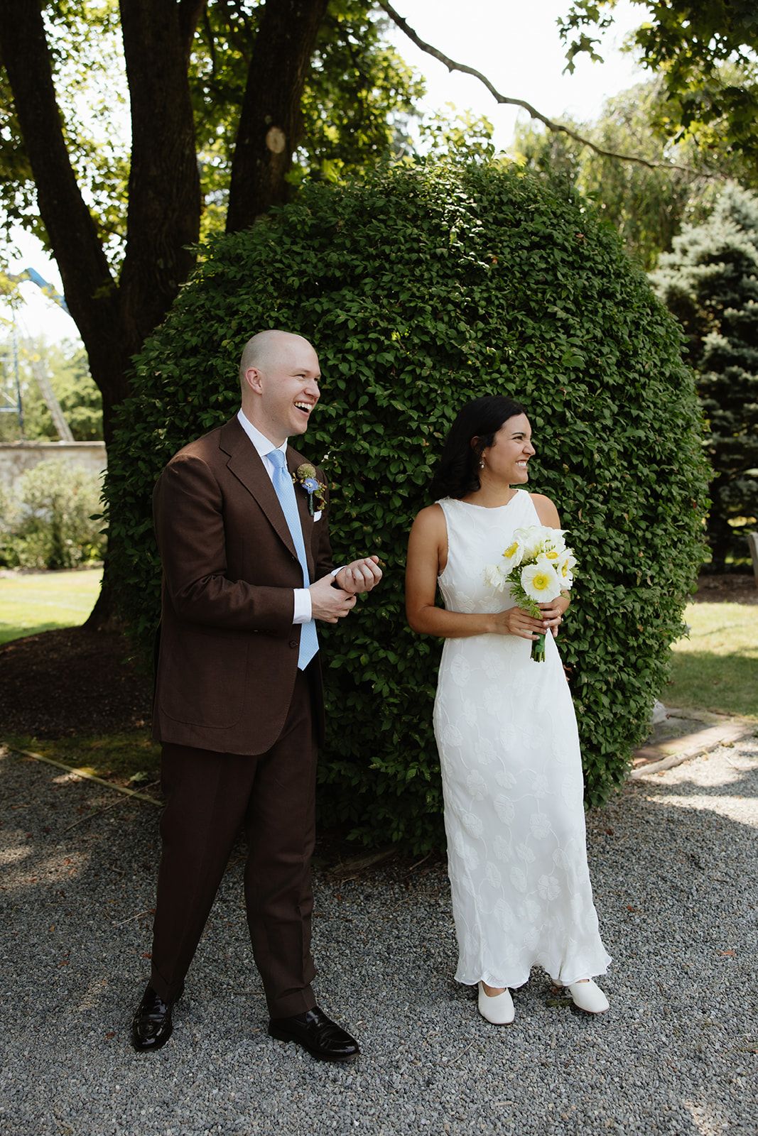 A wedding couple shares a playful moment on a gravel path next to a lush green hedge in a garden setting.