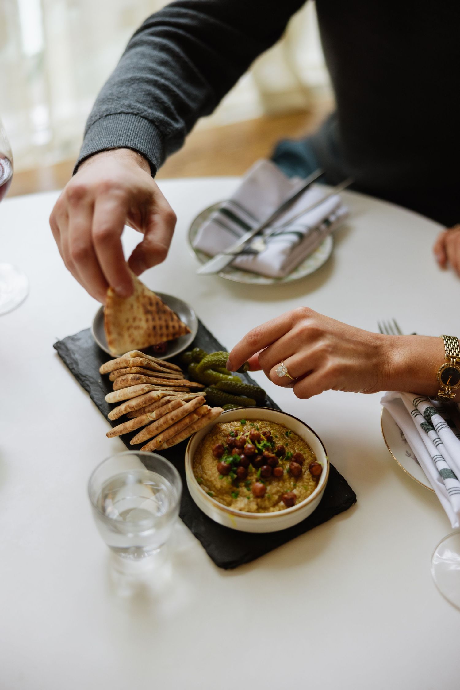 Hands reaching for appetizers served on slate board with hummus dip and pita bread at restaurant table.
