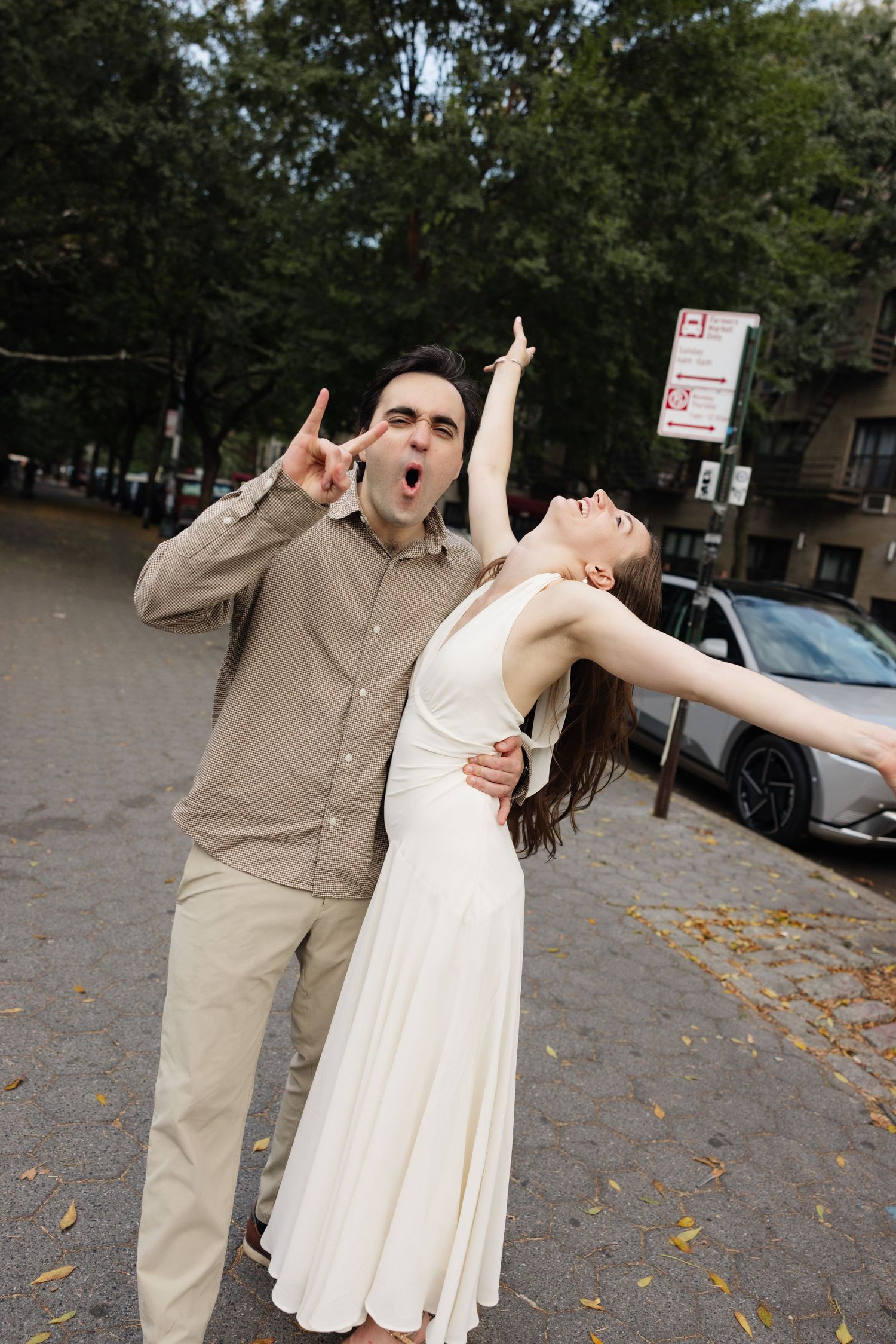 A couple in casual attire dances playfully in a parking lot with fallen autumn leaves scattered on the ground.