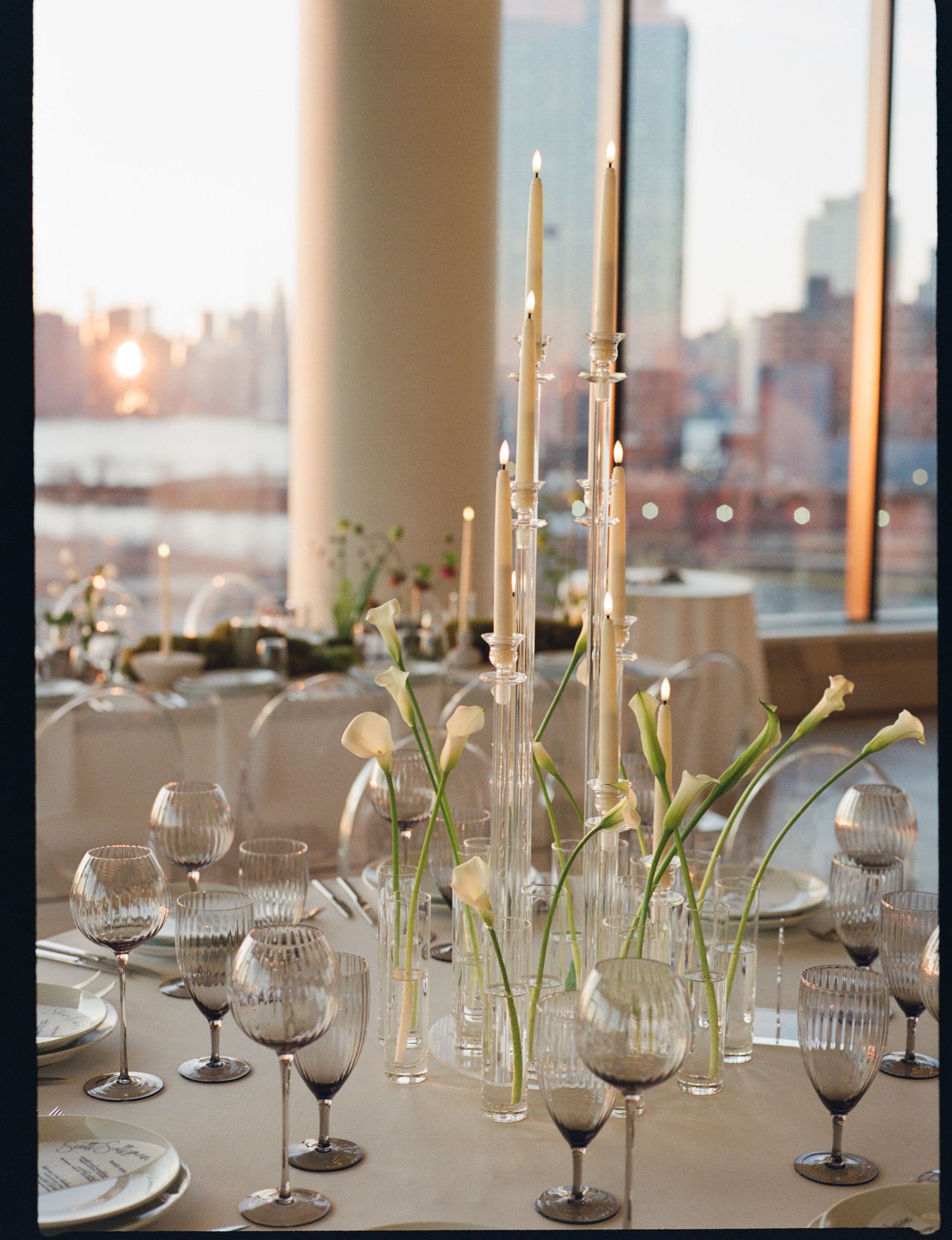 Elegant wedding table with tall taper candles, calla lilies in glass vases, and ribbed stemware overlooking a city skyline.
