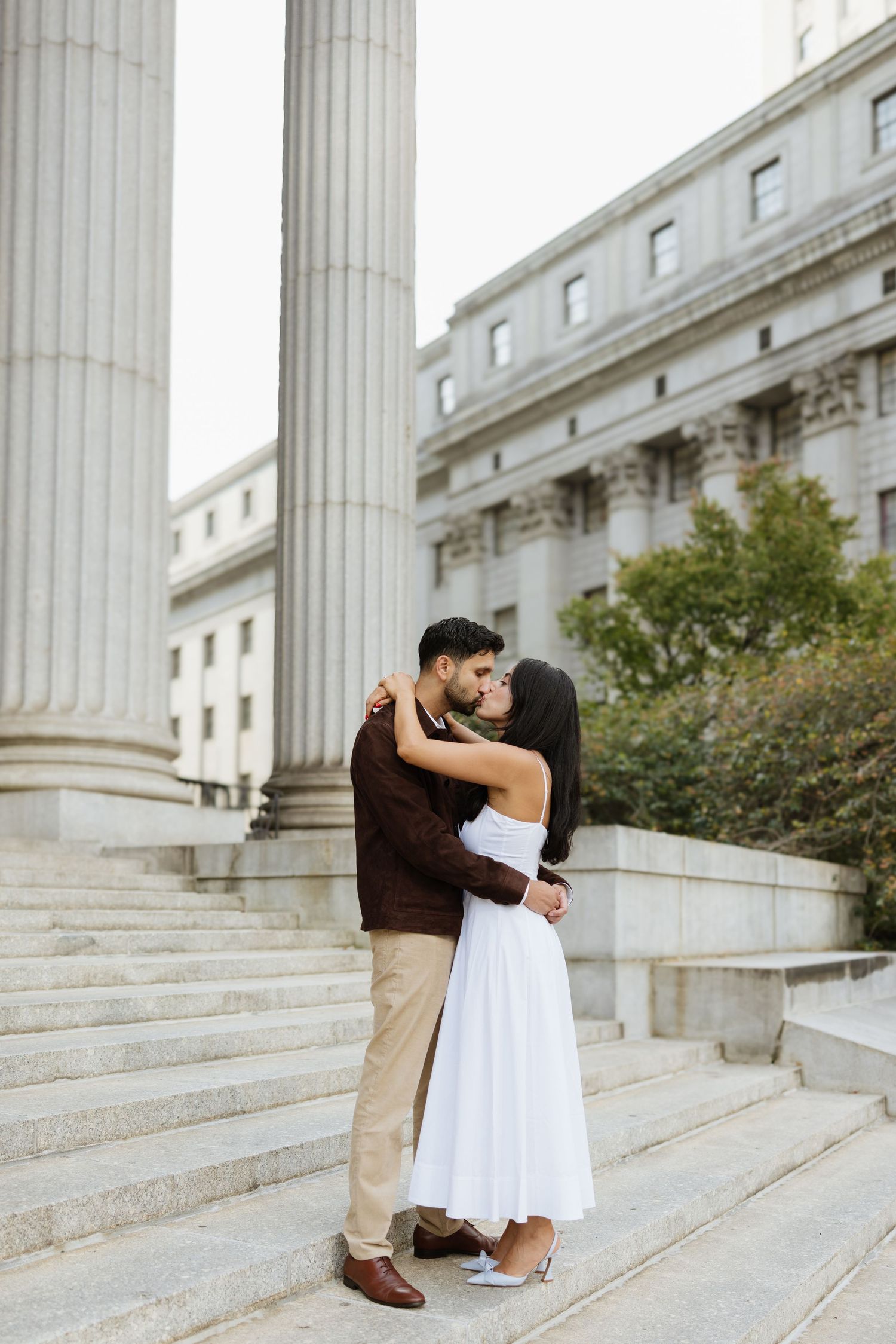 A couple shares a romantic moment on white marble steps outside a classical building with columns.