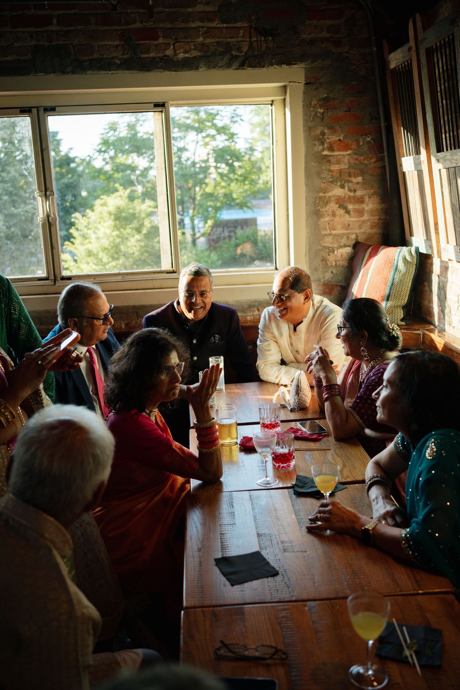 Group of people in traditional Indian attire gathered around a wooden table, sharing drinks and conversation near a brick wall window.