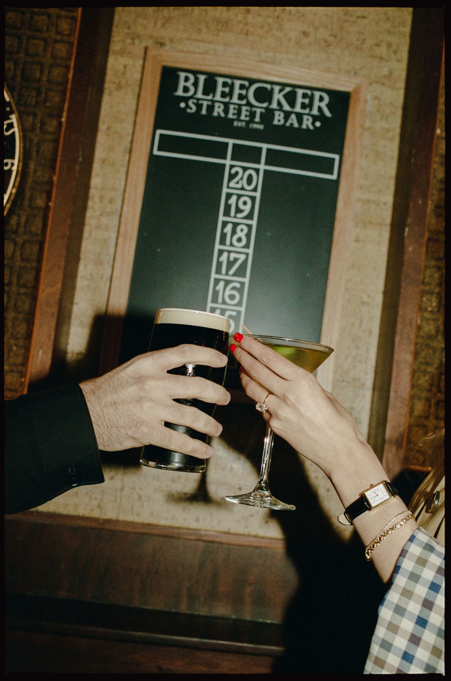 Two people toasting drinks at Bleecker Street Bar, one holding a dark beer and the other a martini glass.