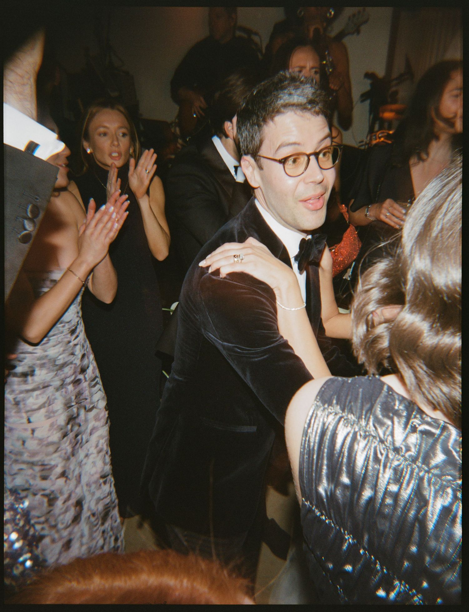 Man in black tuxedo with glasses dancing at elegant formal party event surrounded by guests in evening wear.