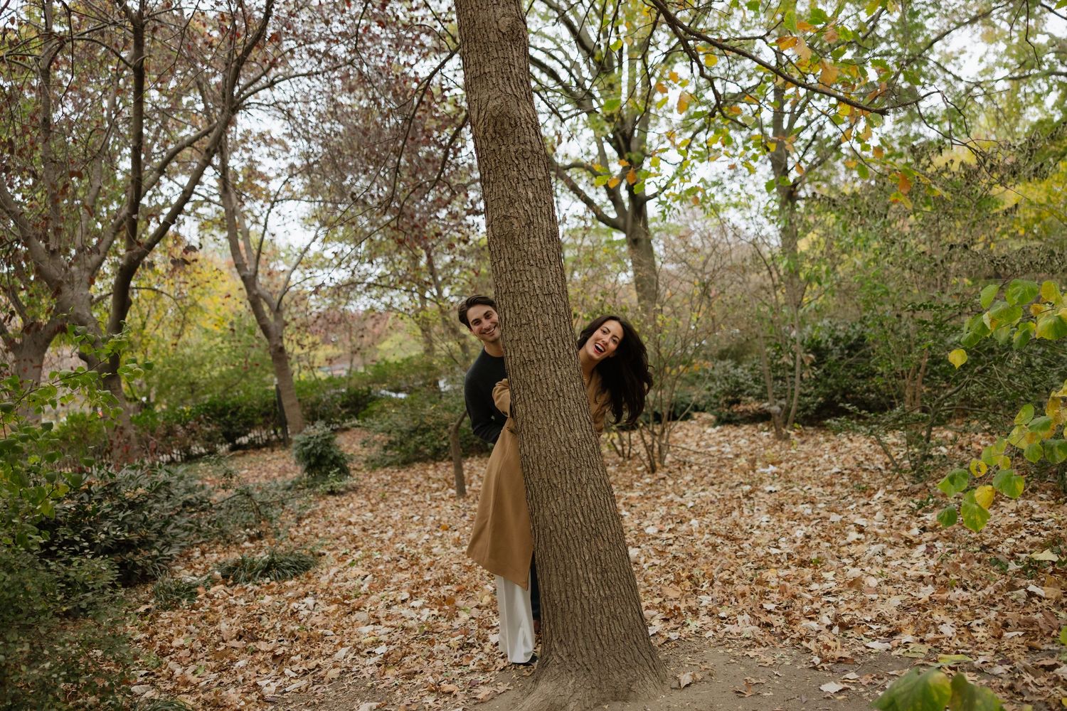 Two people in brown coats pose playfully on opposite sides of a tree trunk in an autumn forest setting.