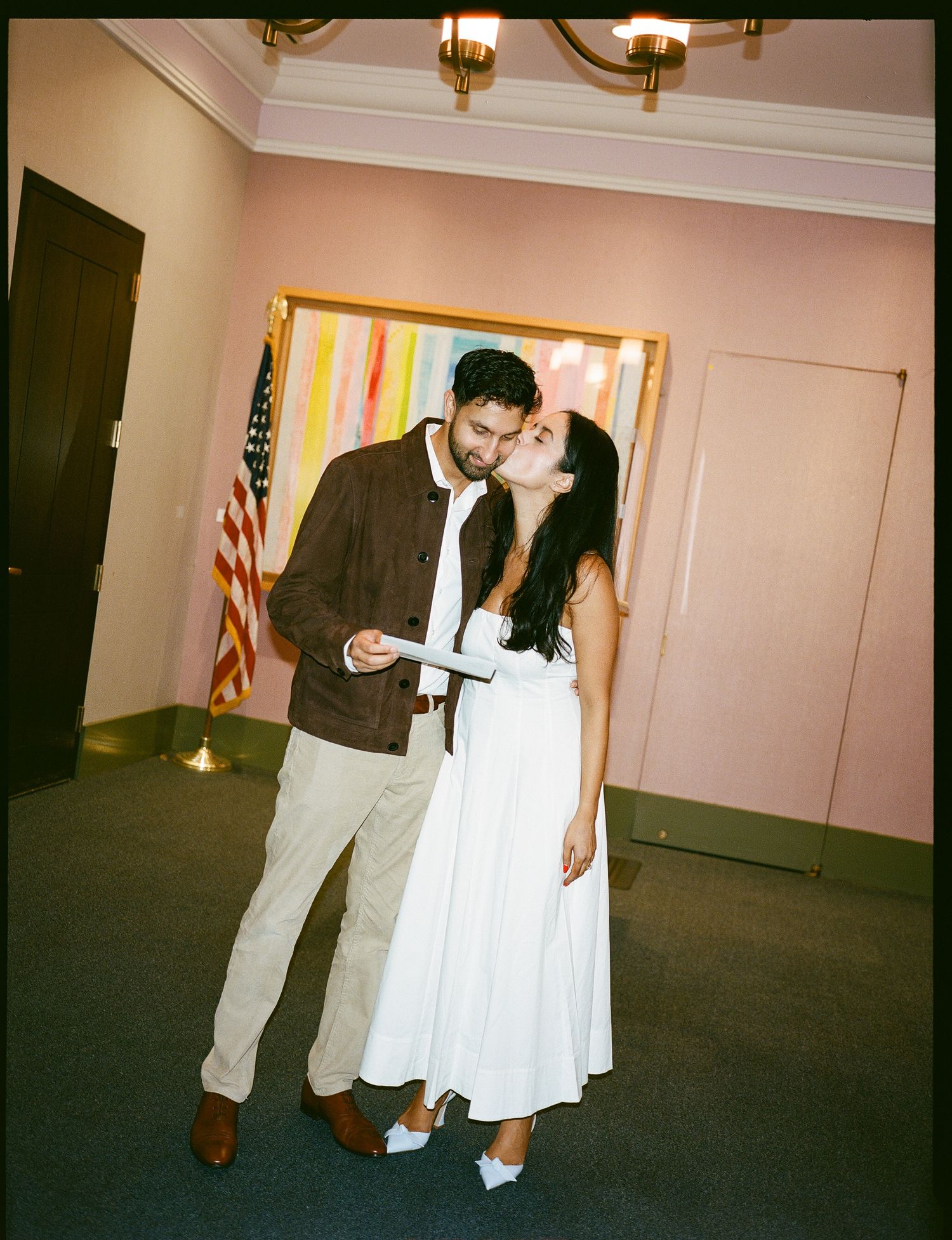 A couple in formal attire dance together in a room with soft lighting and an American flag in the background.