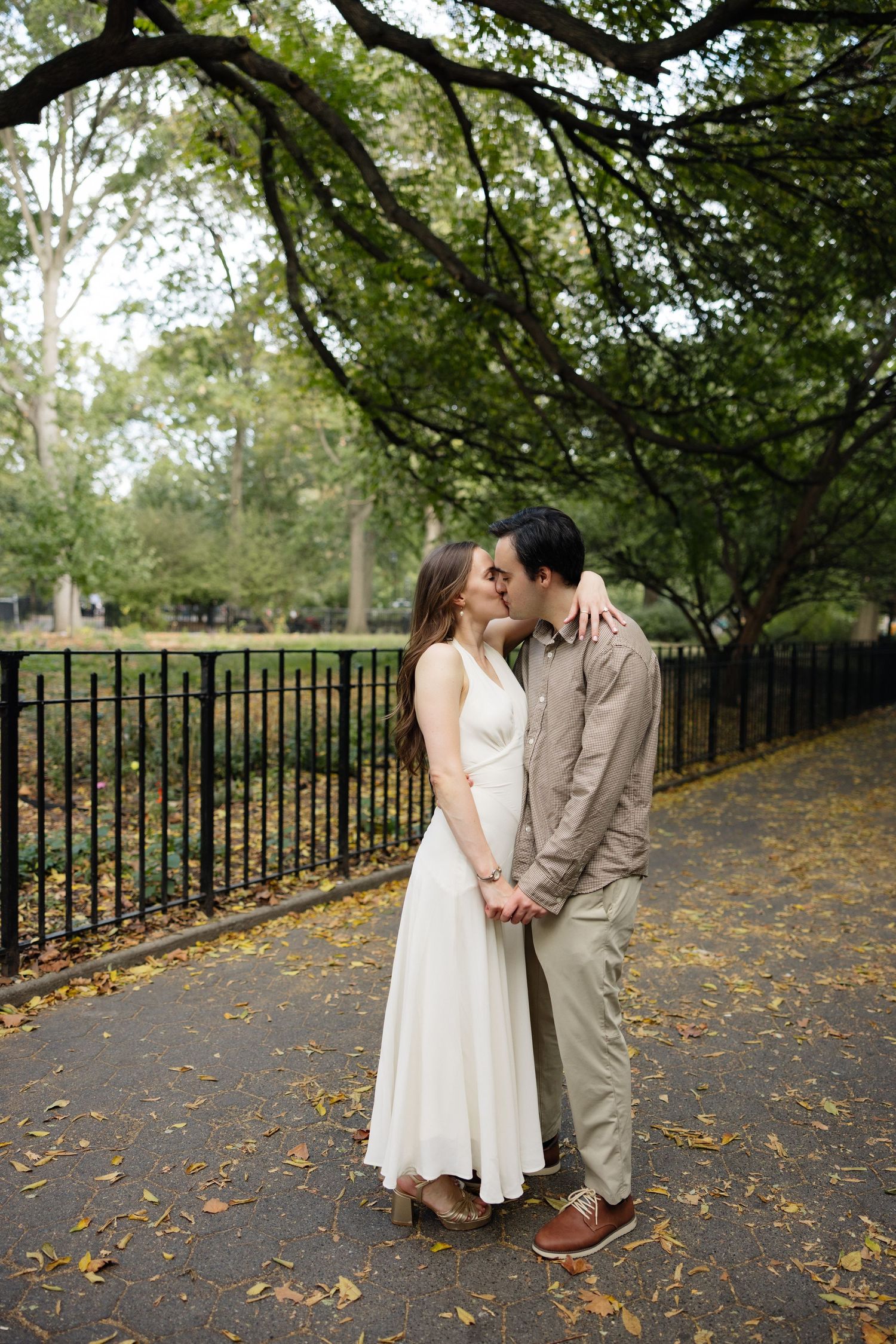 A couple shares a romantic moment on a tree-lined path covered in fallen autumn leaves.