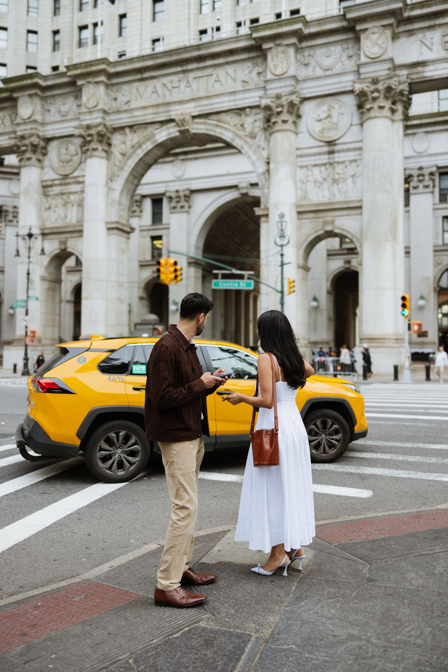 Two people stand on a street corner in front of classic New York architecture with a yellow taxi cab passing by.