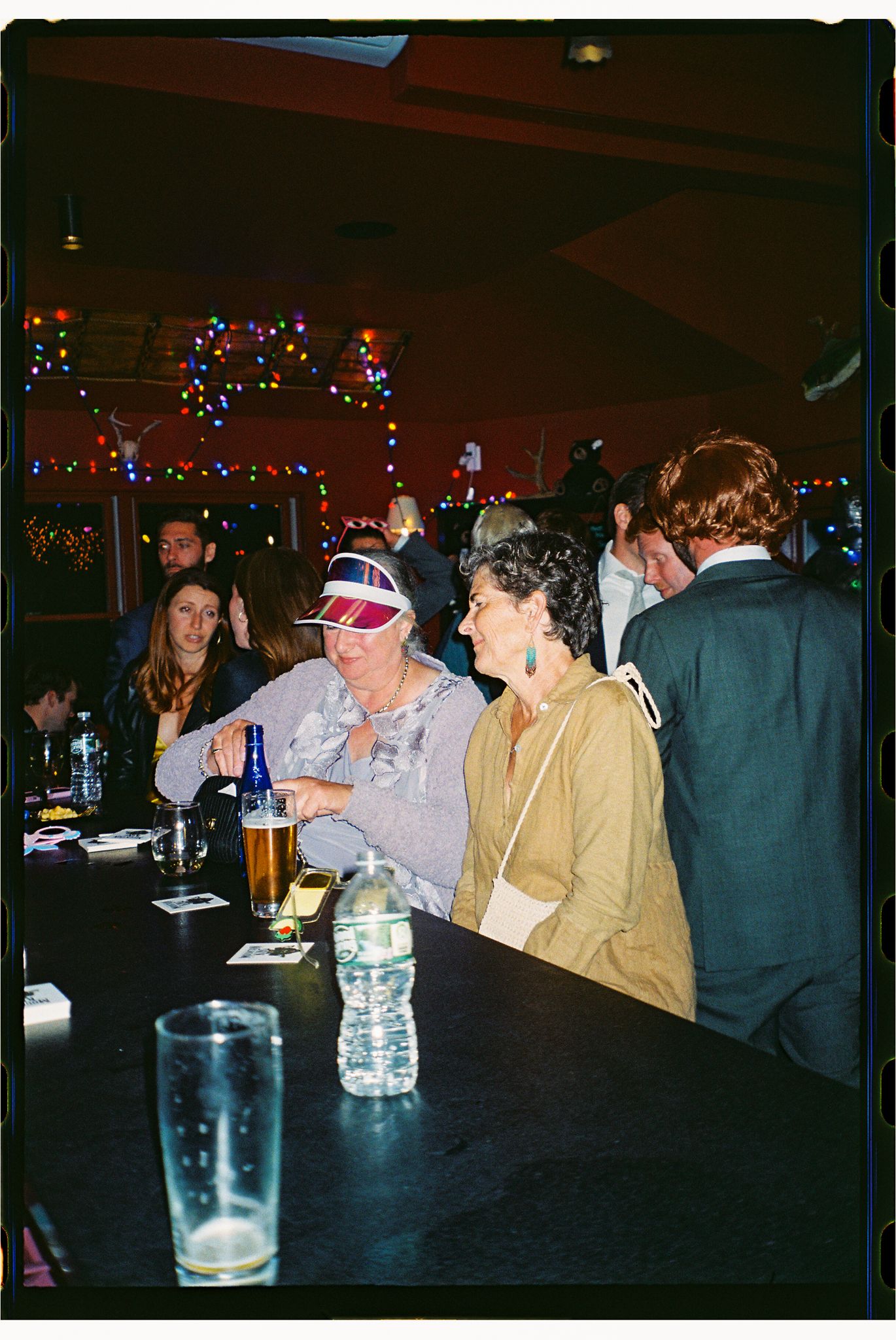 A dimly lit indoor event space with people seated at tables decorated with colorful string lights and water bottles.