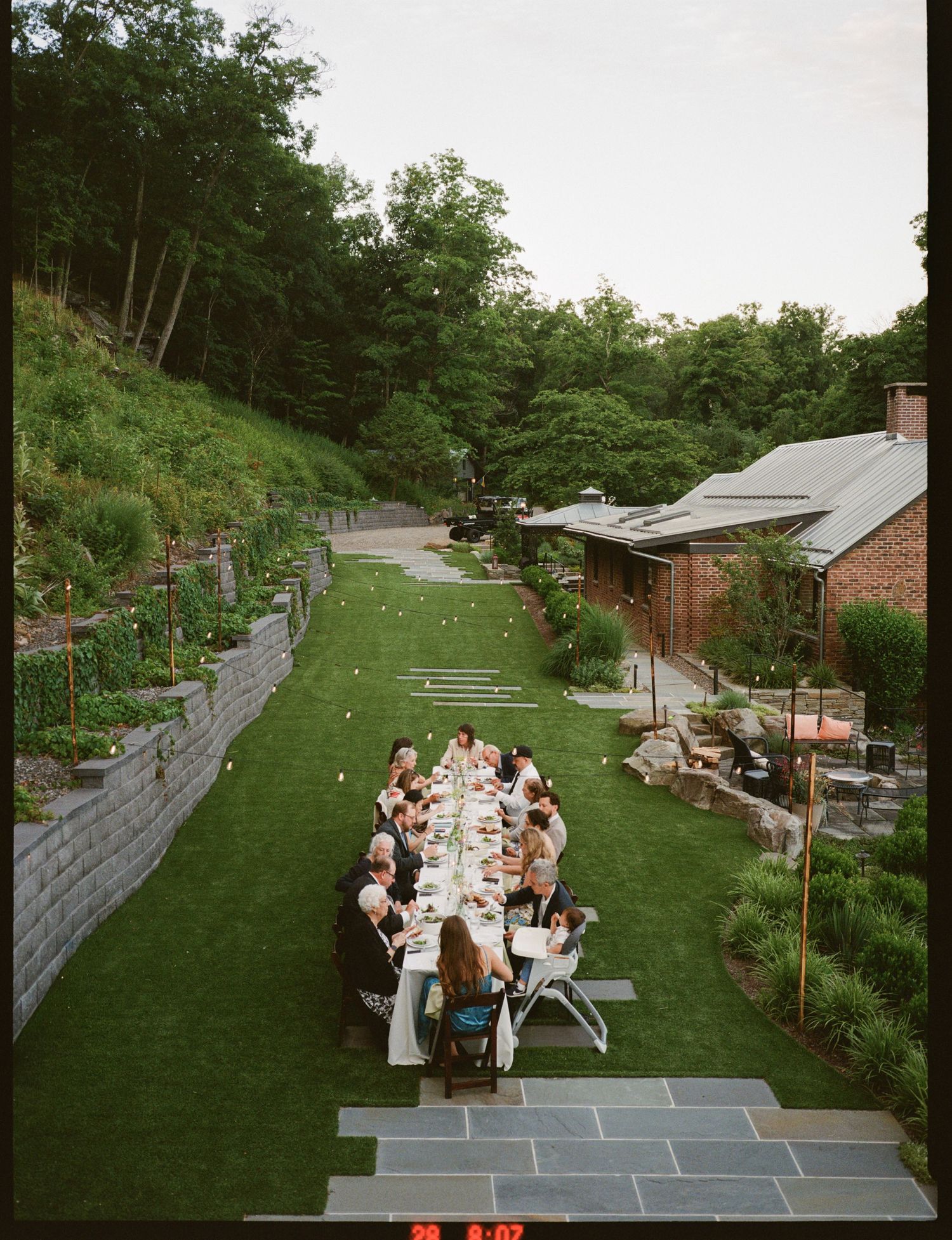 Aerial view of an elegant outdoor garden dinner party with guests seated at a long table on lush green lawn.