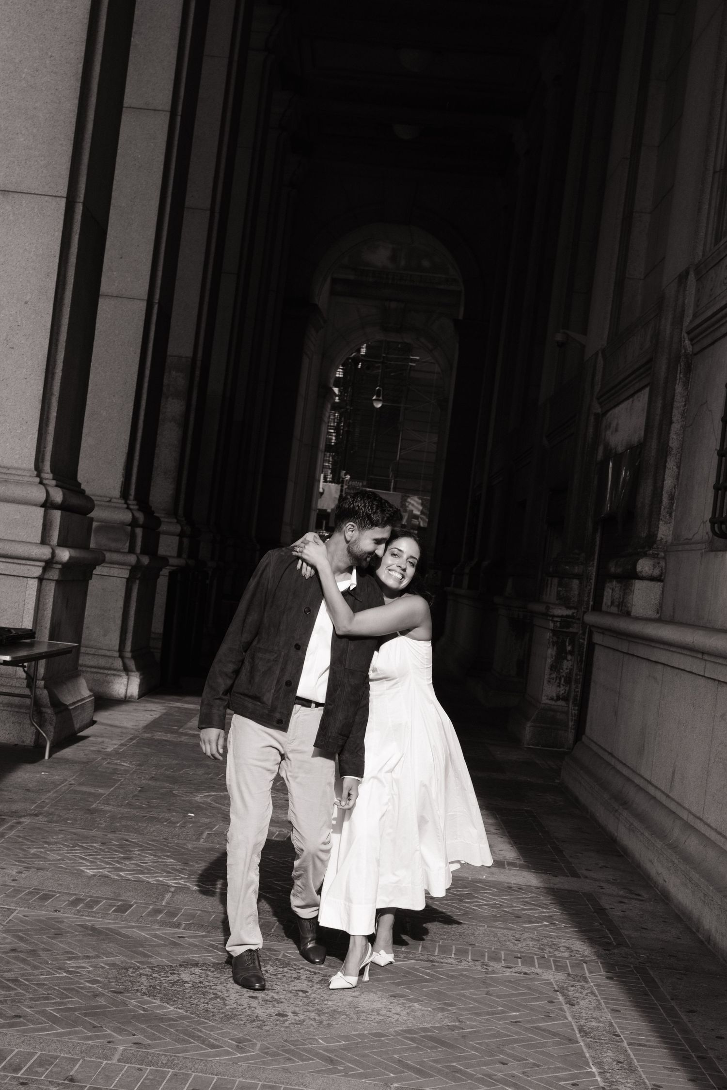 A romantic black and white photograph of a couple embracing in an elegant arched corridor with stone columns.