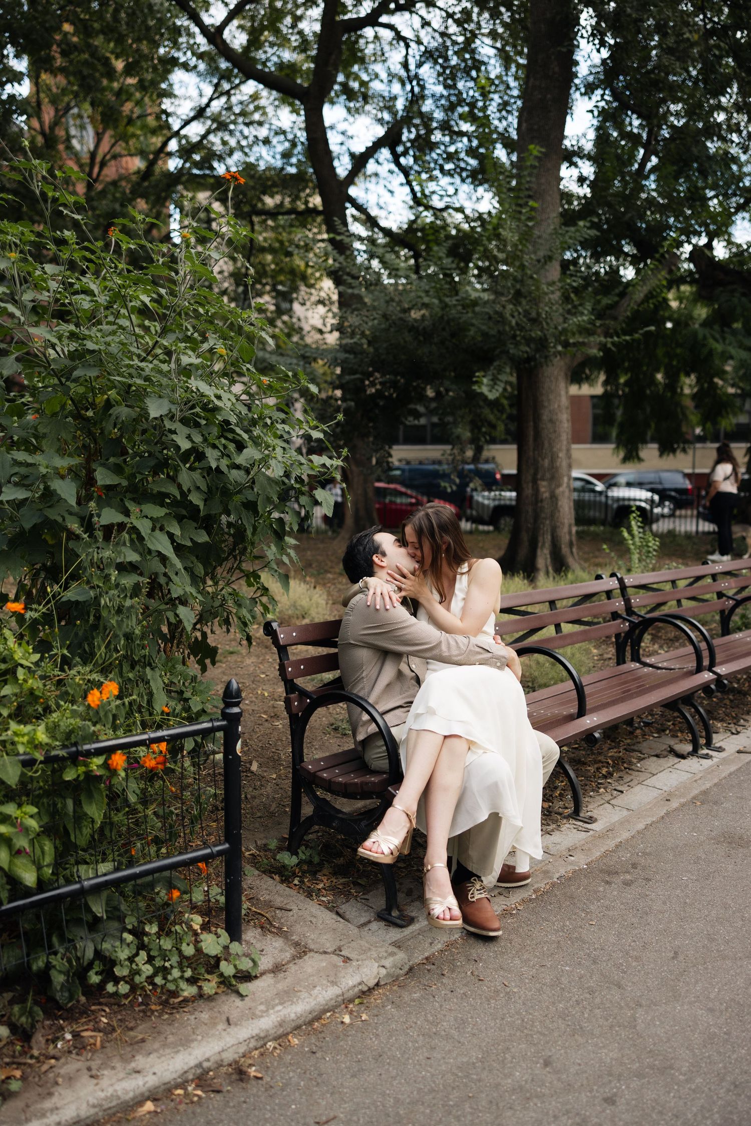 A couple sits close together on a wooden park bench surrounded by lush greenery and flowers on a summer day.