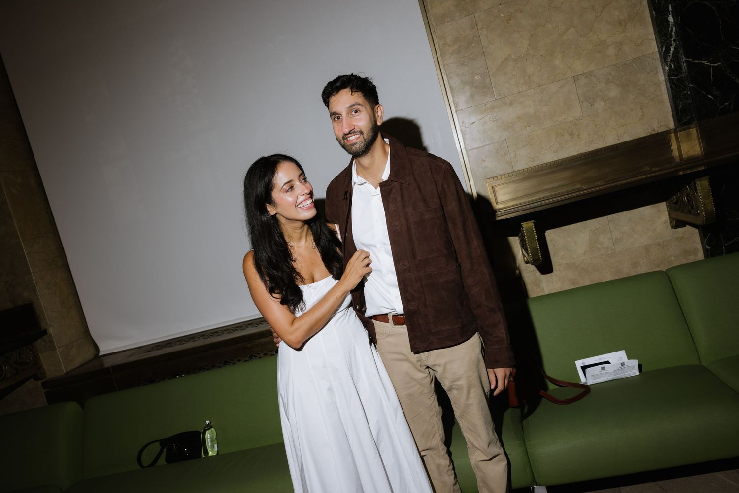 A couple in casual evening wear share a candid moment together while standing near a green couch.