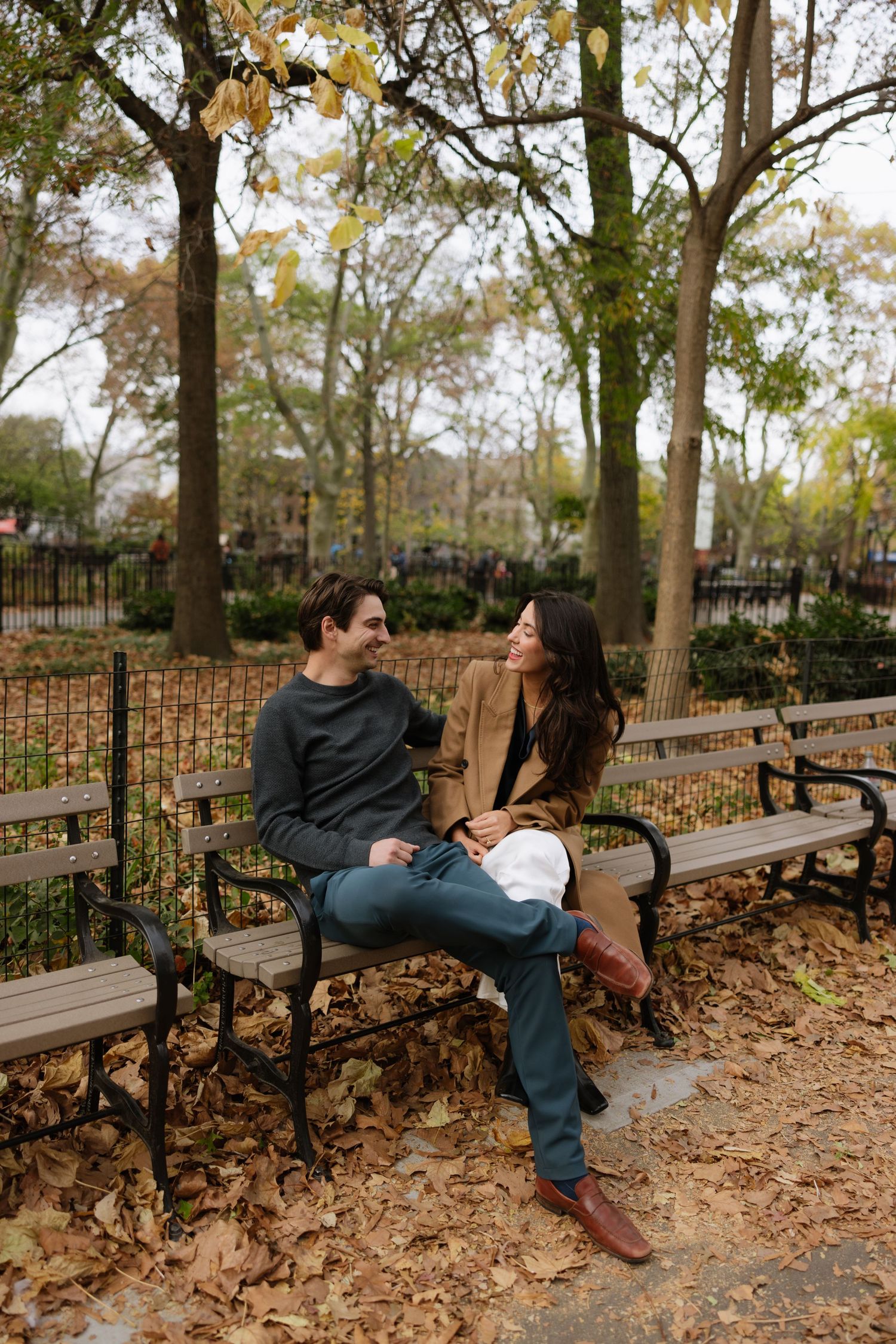 A couple sits together on a park bench surrounded by autumn leaves and trees in a peaceful outdoor setting.