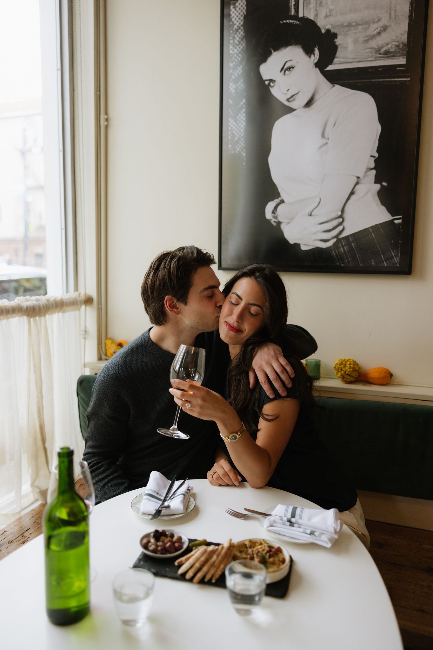 A romantic couple shares an intimate moment at a restaurant table with wine glasses and plates.