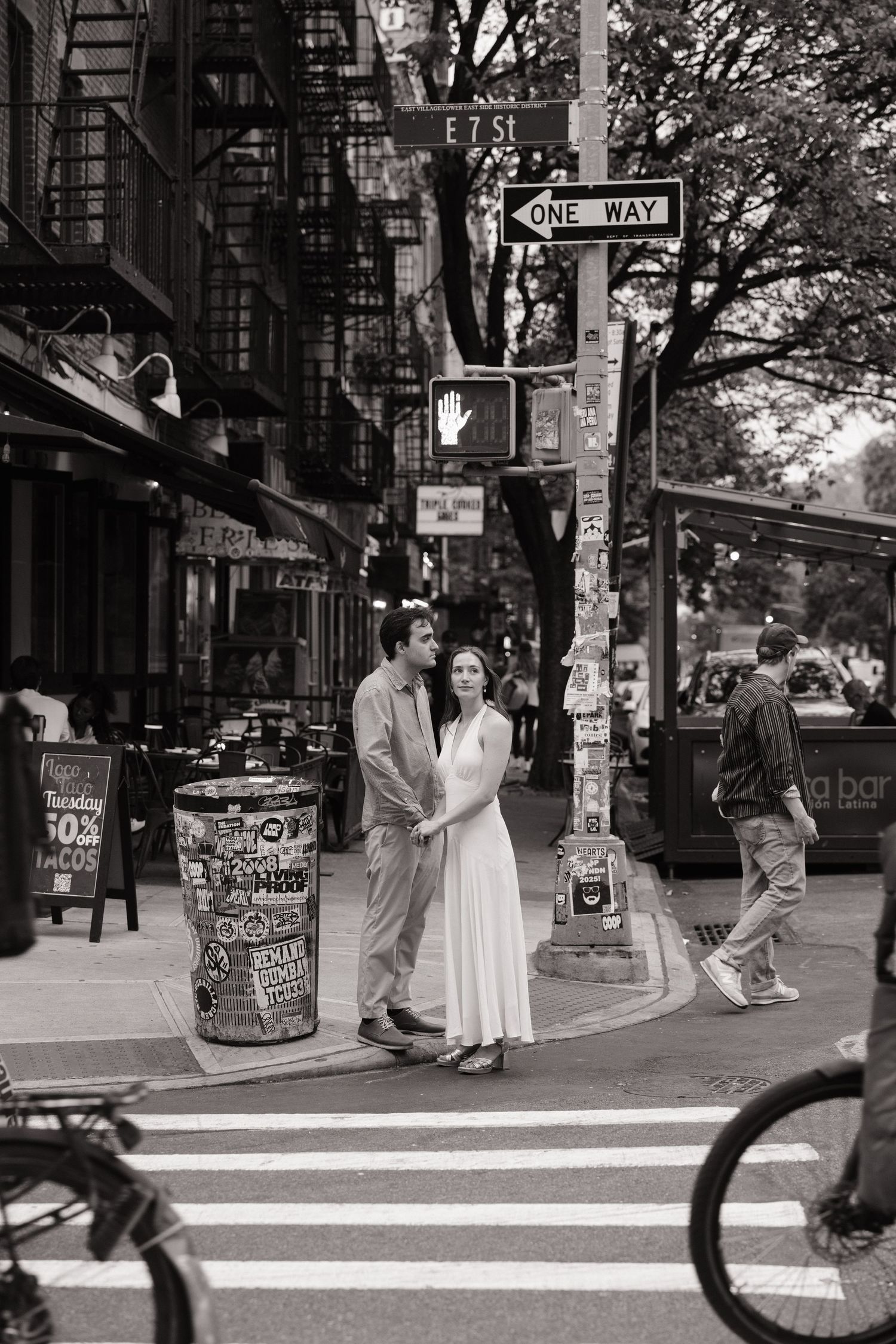 Black and white street photography capturing people interacting on a city sidewalk with posters on poles.