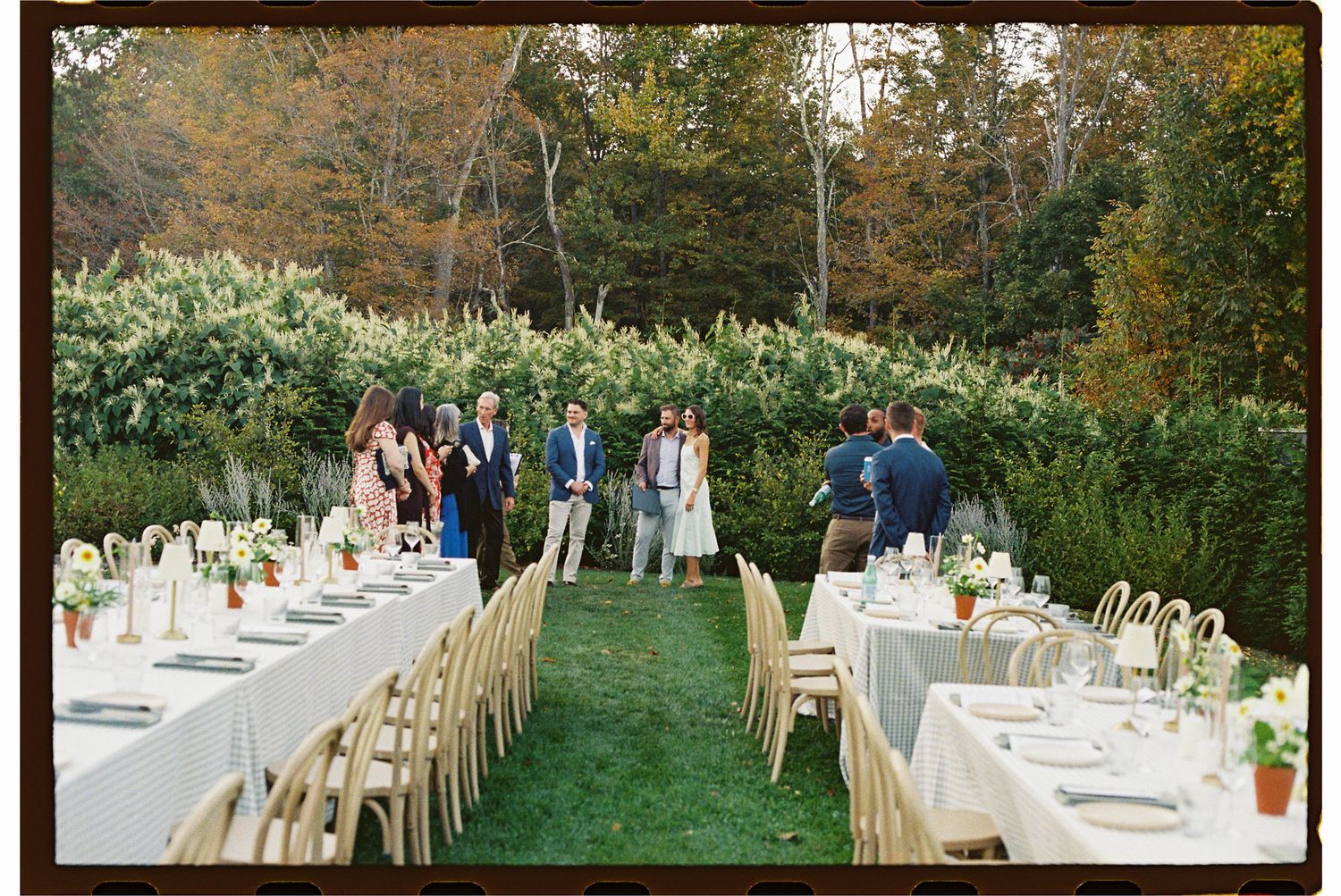 Long wooden tables with white linens are set up outdoors for an elegant garden wedding reception under autumn trees.
