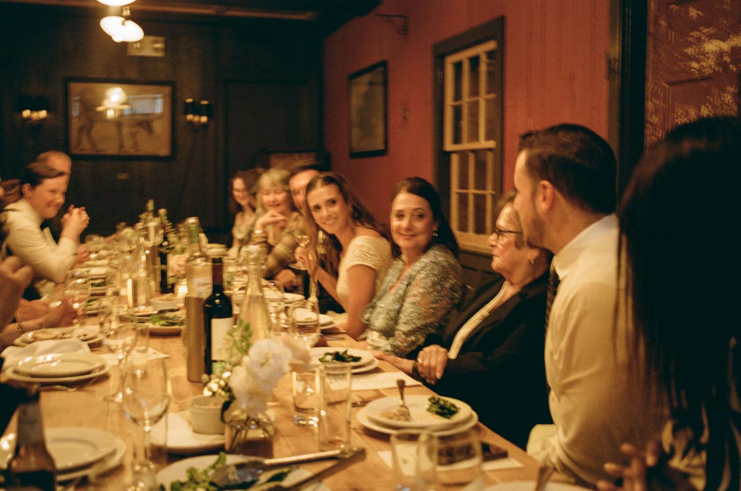 Guests enjoy a candlelit dinner party at a long rustic table in a warmly lit red dining room.