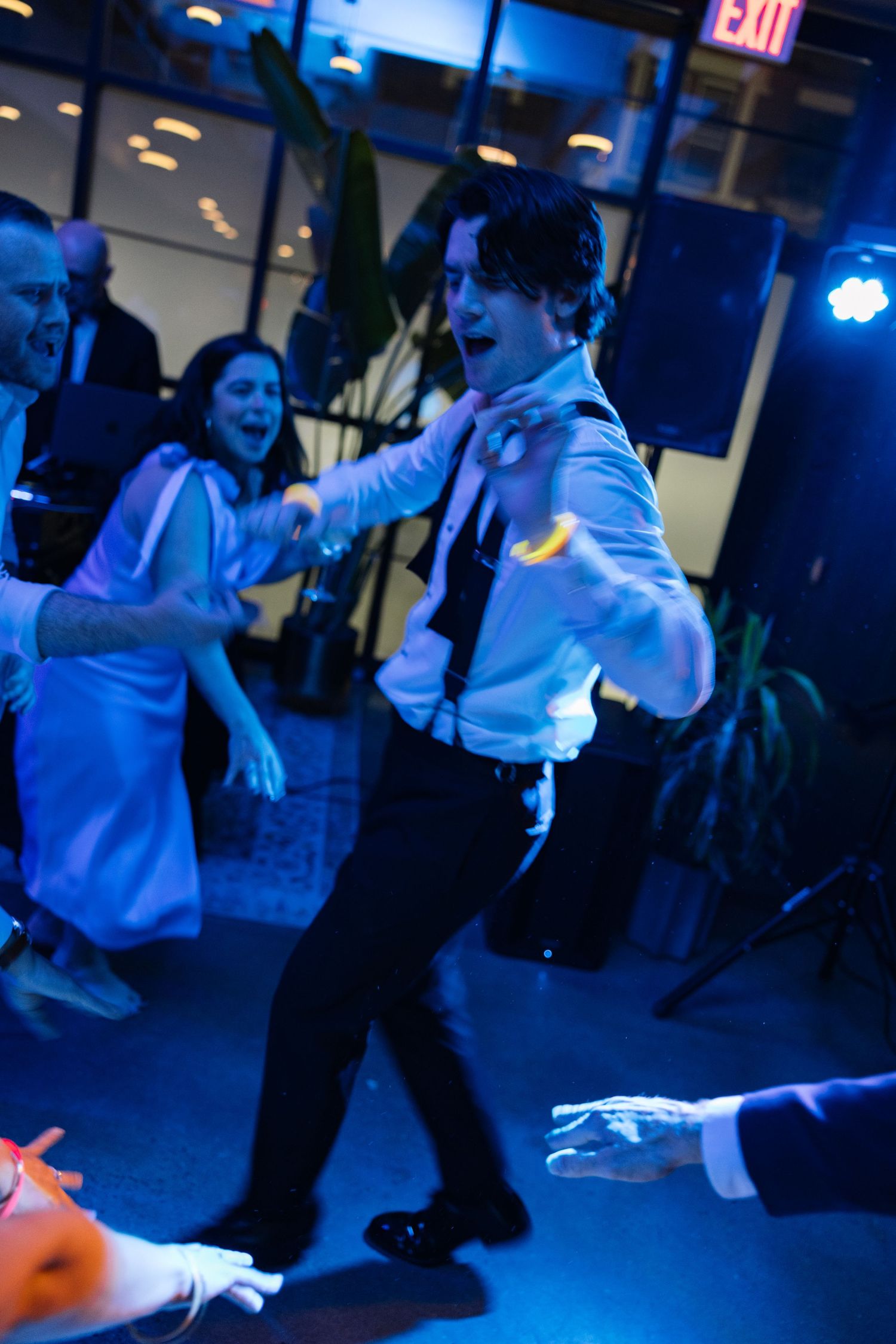 Young man dancing energetically at a wedding reception under blue lights, wearing white shirt and black suspenders.