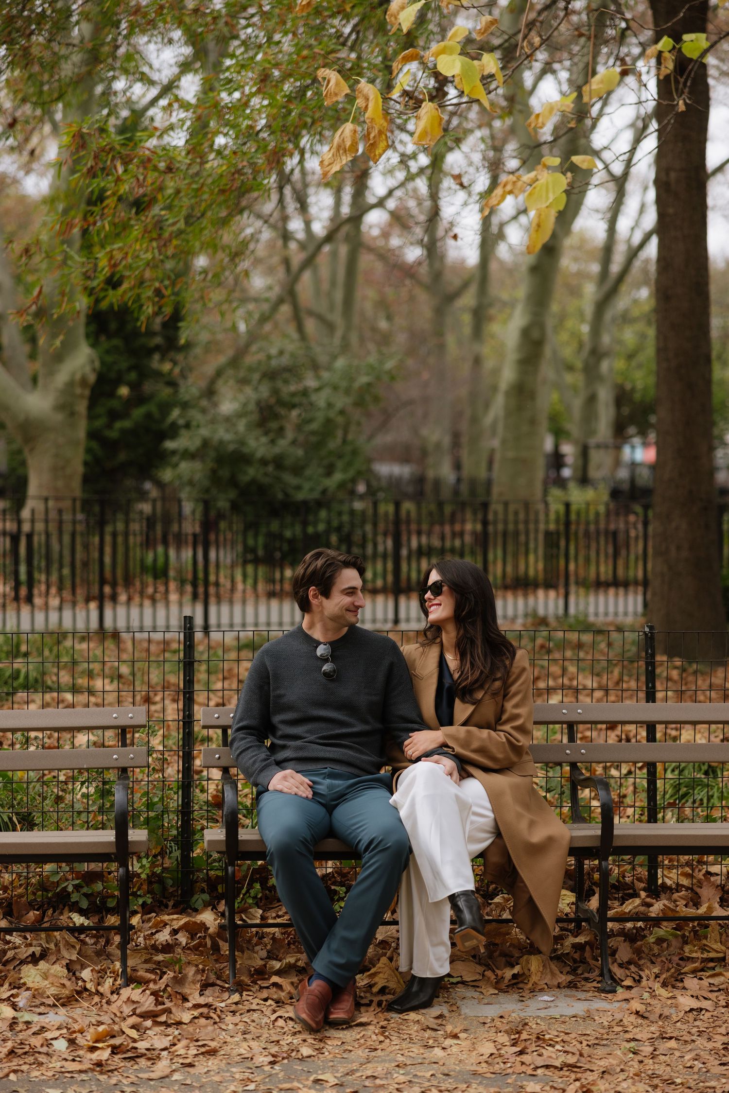 A couple sits together on a park bench surrounded by fallen autumn leaves and an iron fence.