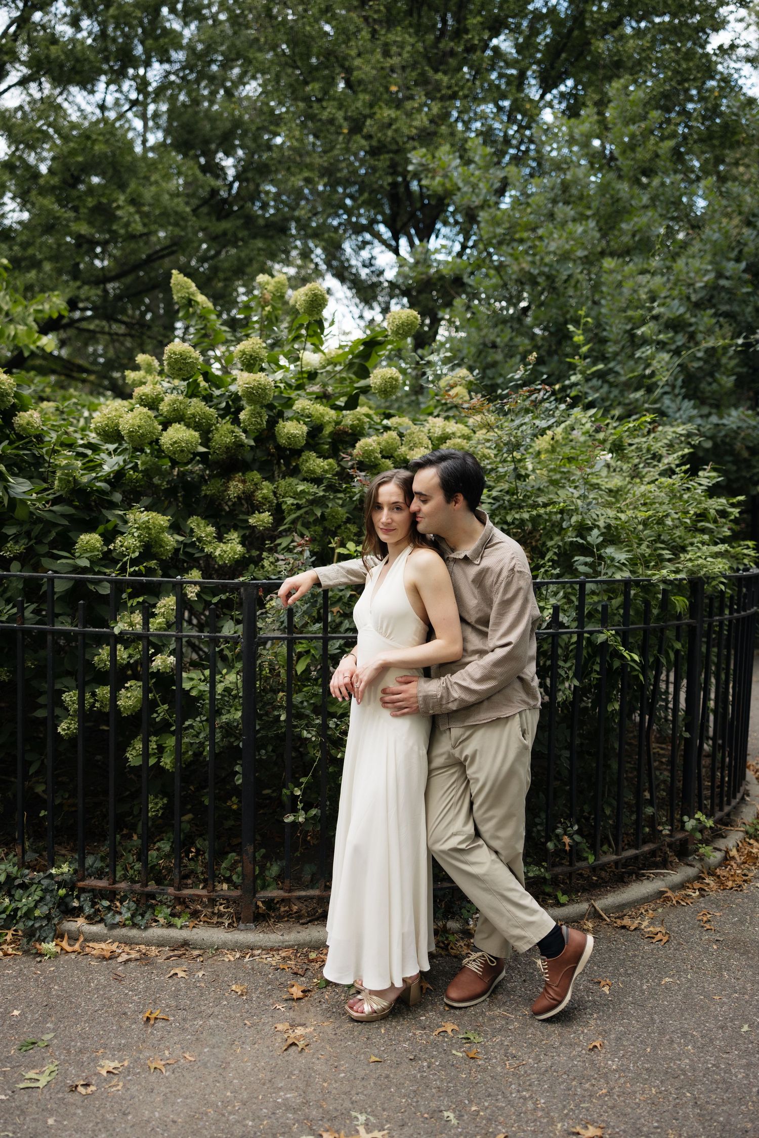 A couple in casual attire embraces against an iron railing with lush greenery in the background.
