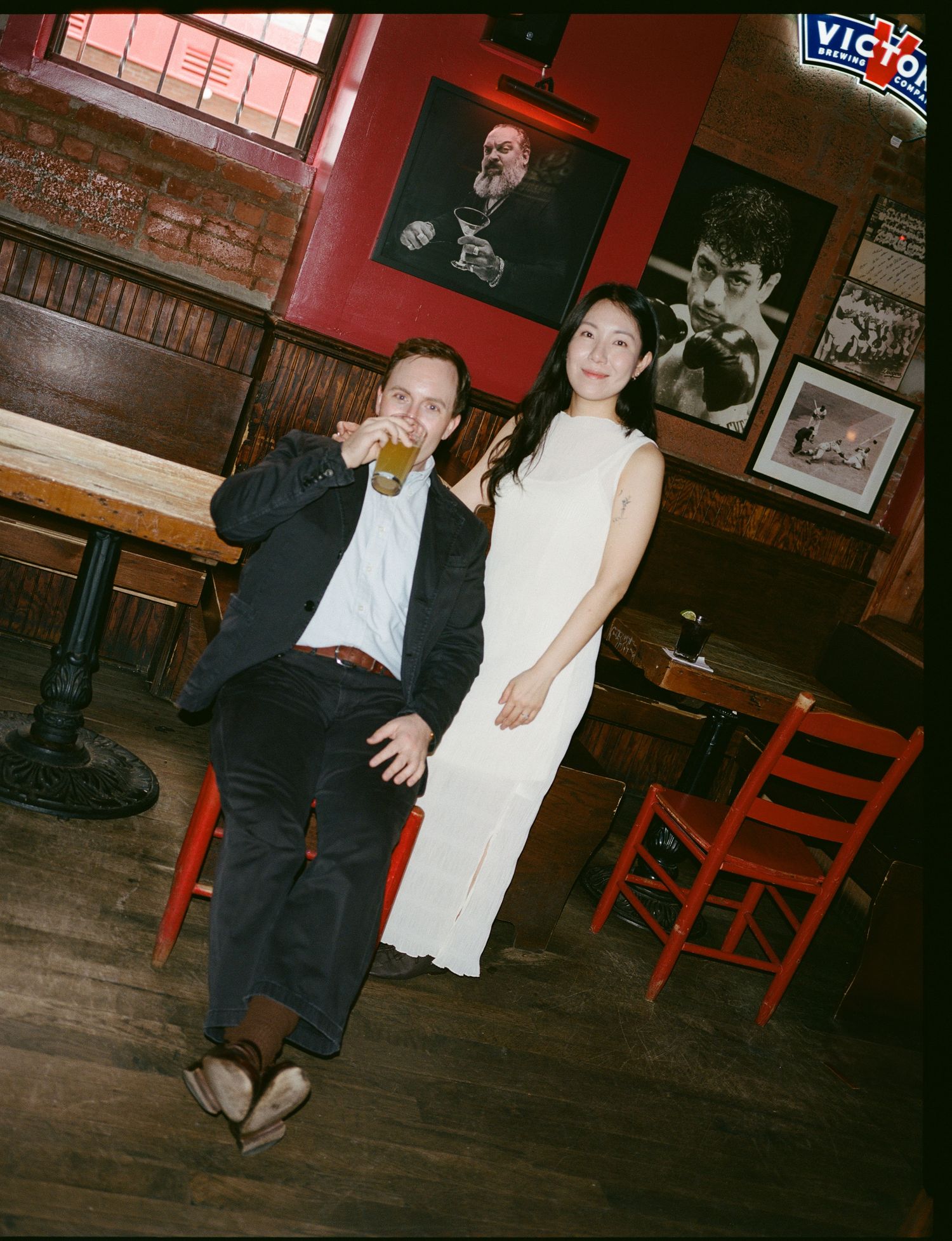 A couple sits together in a dimly lit bar with red walls and vintage photos, as one enjoys a beer.