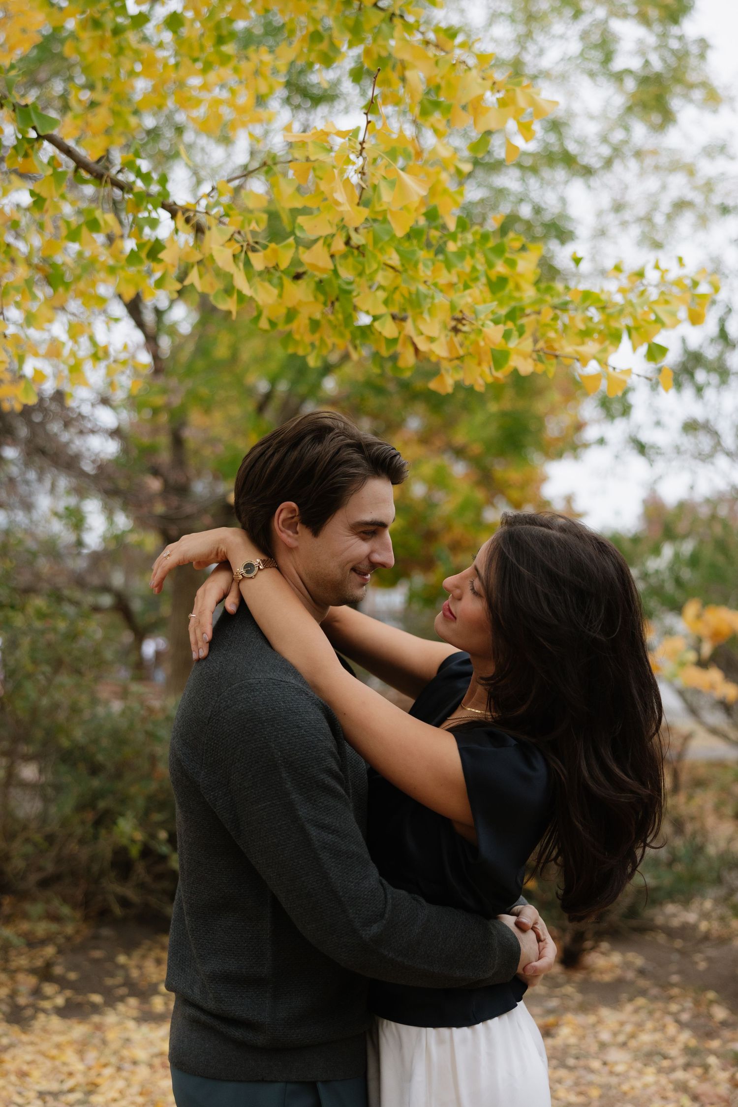 A couple embraces romantically under autumn trees with golden yellow leaves falling in the background.