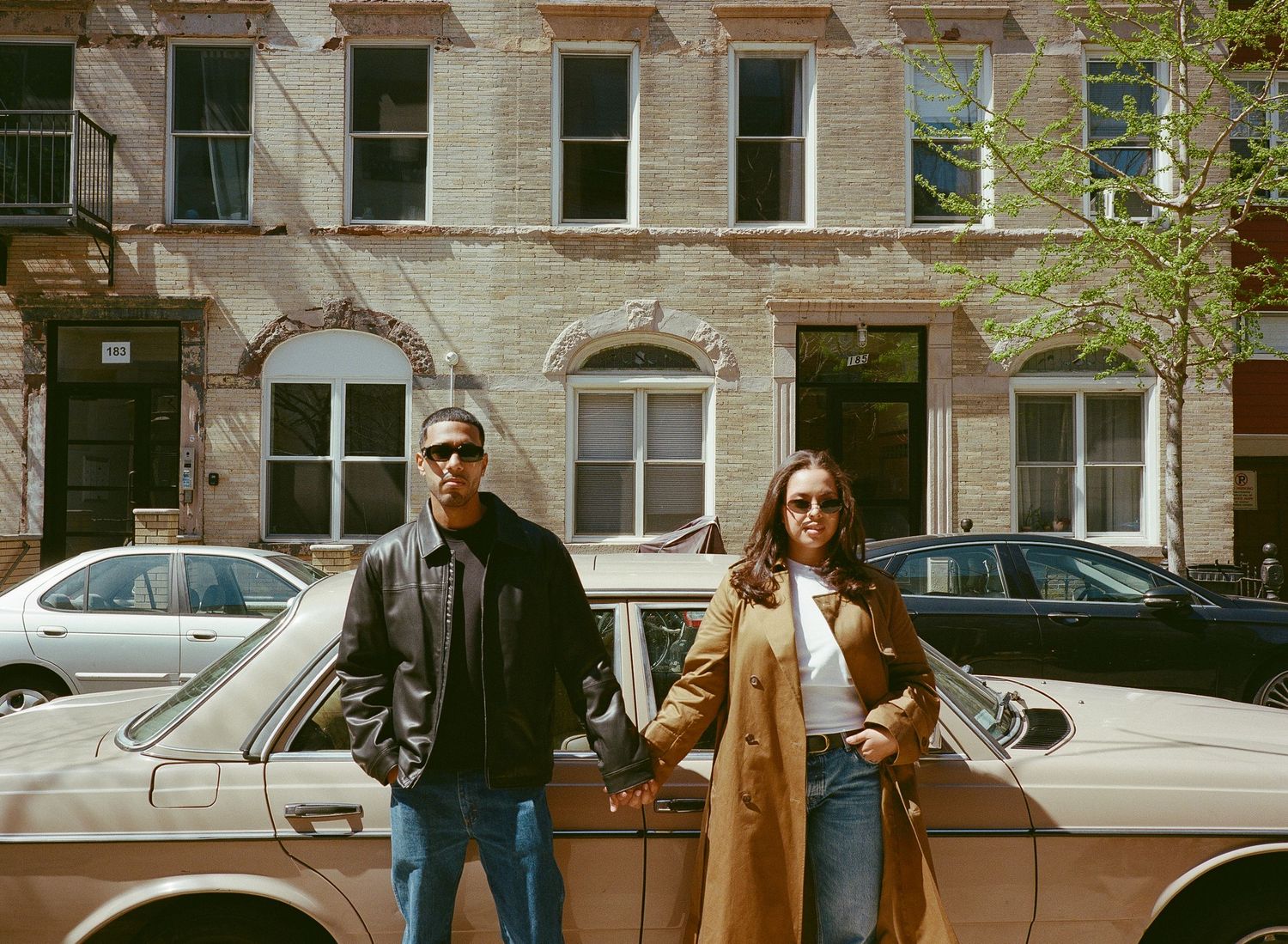 People walking on a city street with brownstone buildings and parked cars on a sunny day.
