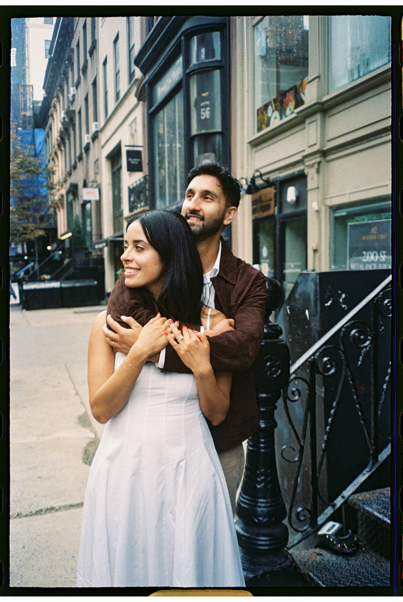 A couple embraces on a charming city street, with the woman in a white dress and the man in a brown jacket.