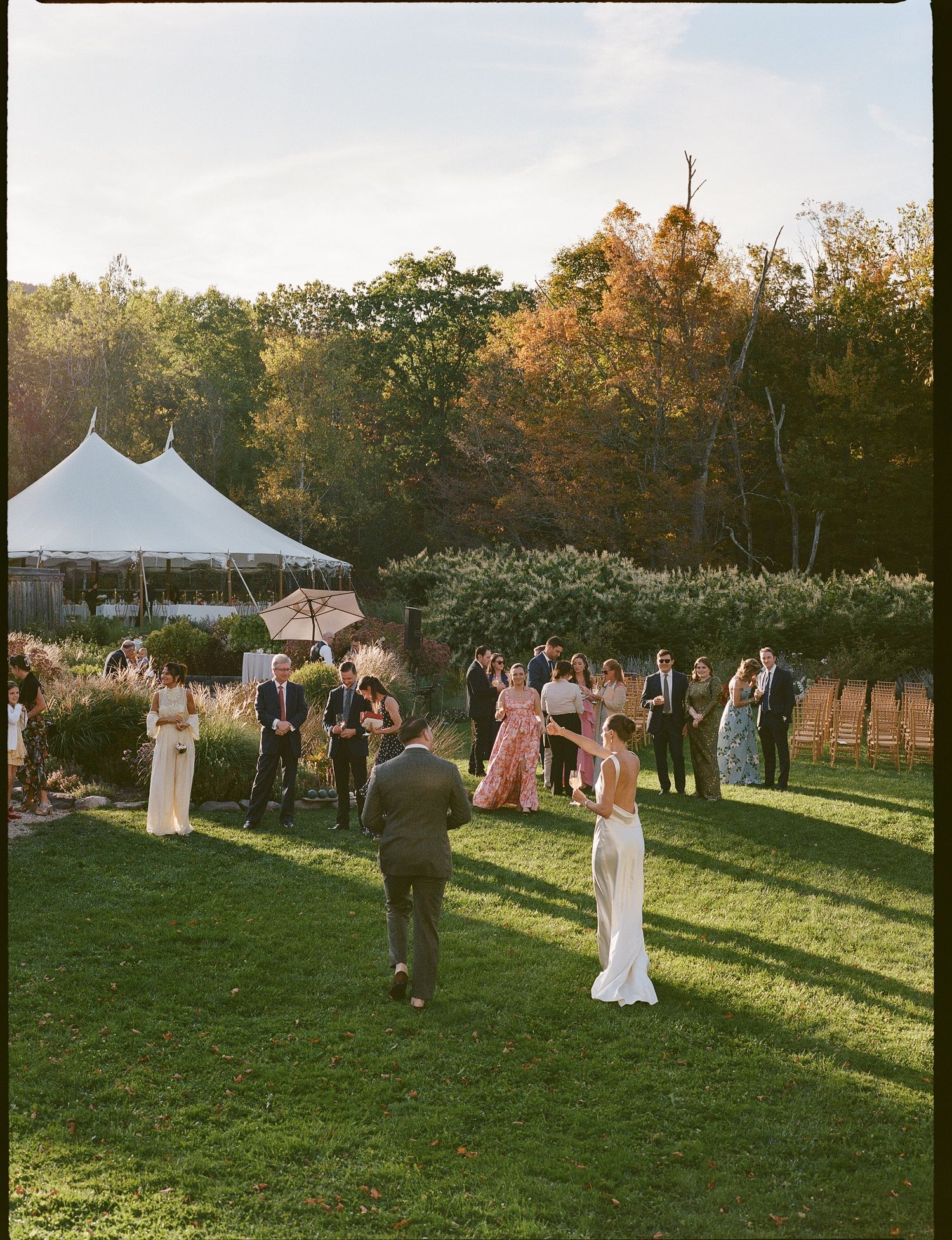 Outdoor wedding reception with white tent and guests mingling on a grassy lawn during golden hour in autumn.