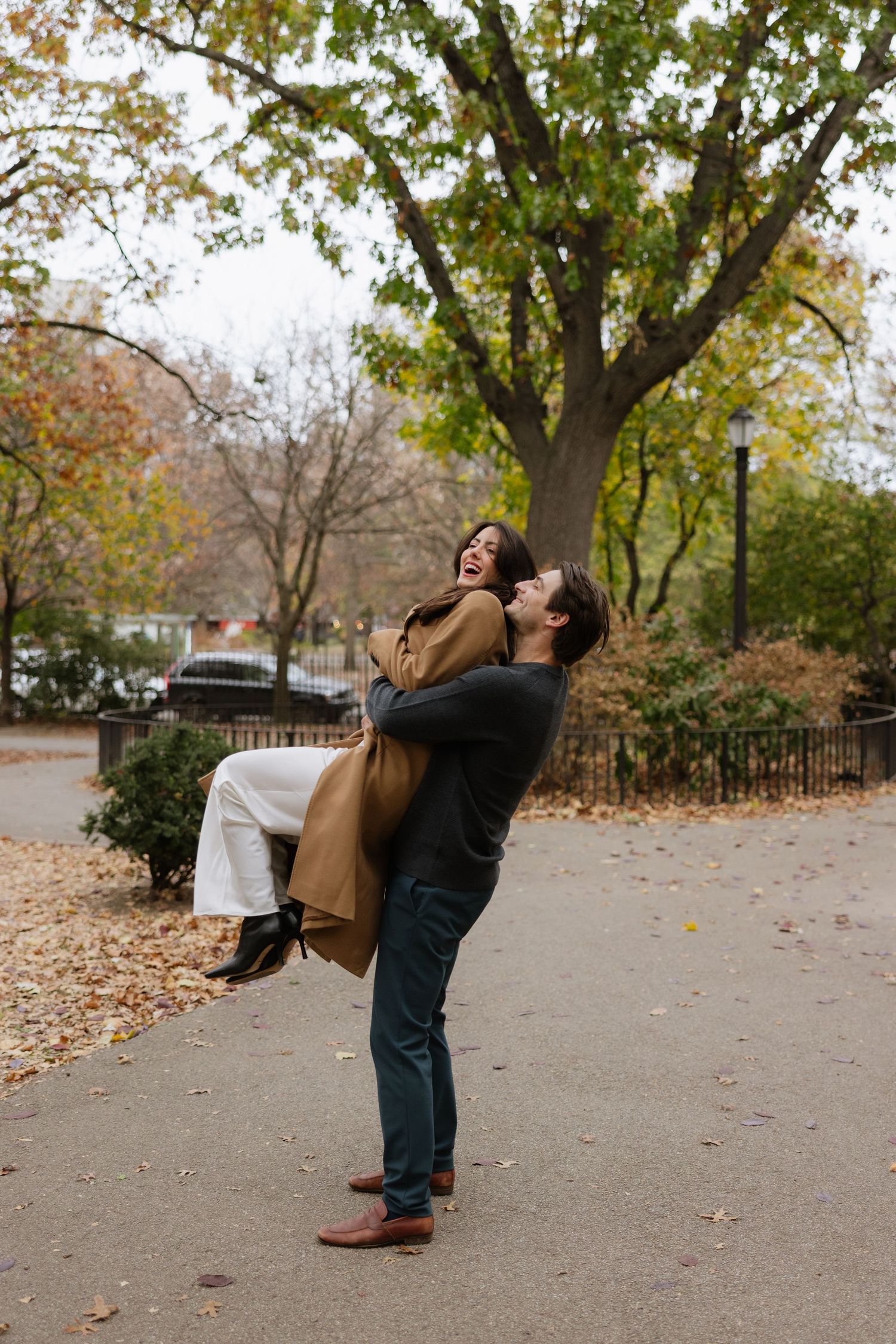 A couple in autumn clothing playfully dances together in a park filled with fallen leaves on a cloudy day.
