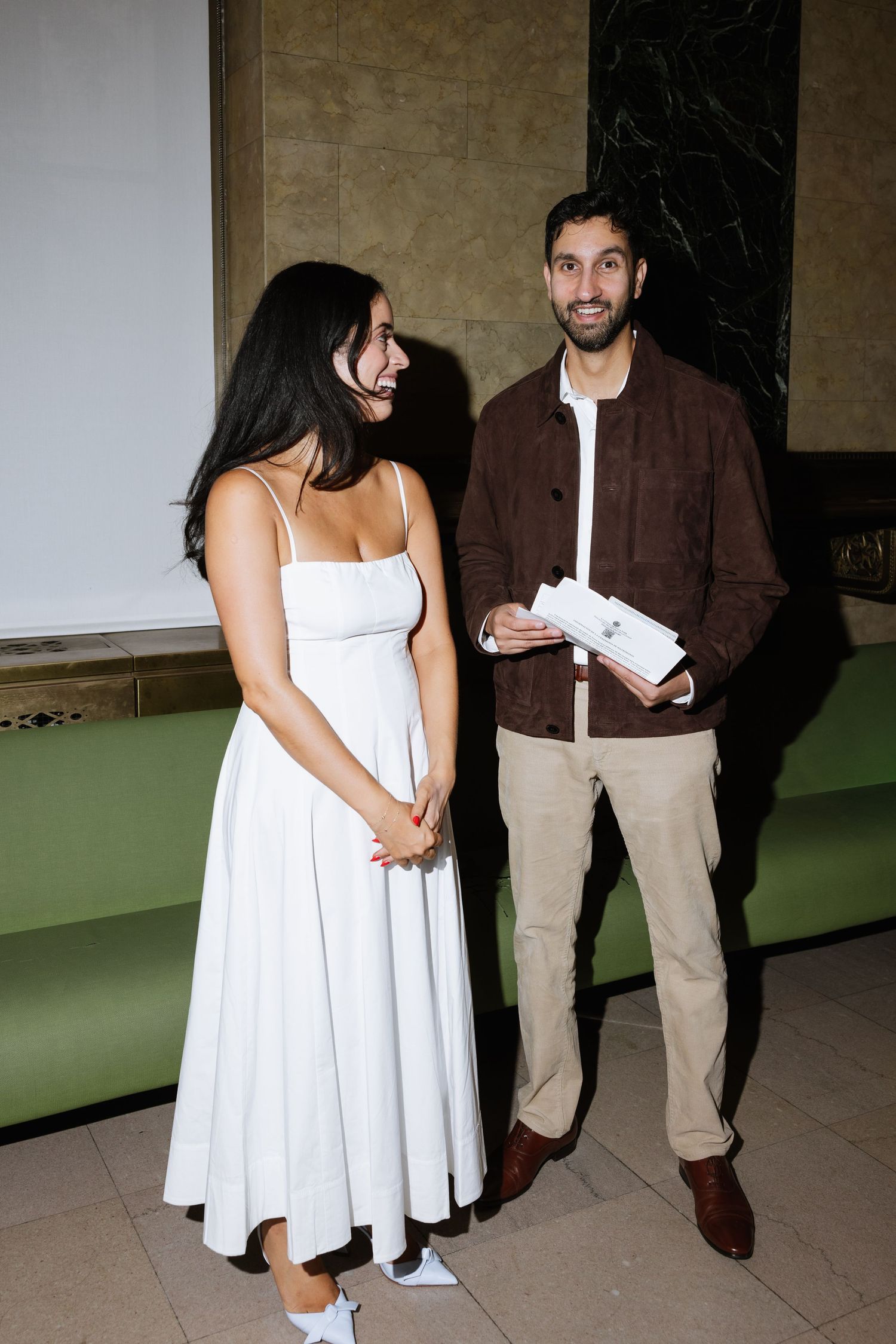 Two people stand together in a hallway, one wearing a white dress and the other in a brown jacket and khaki pants.