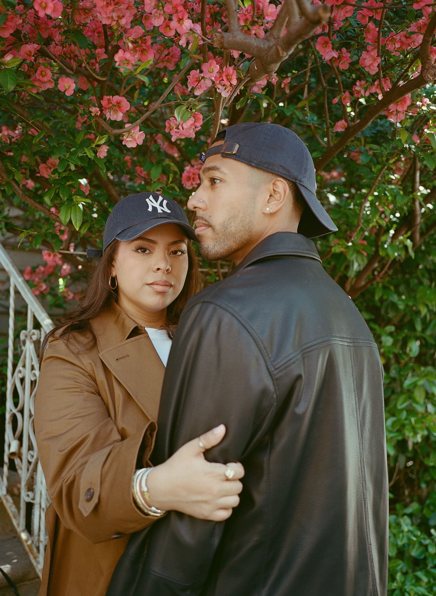 A couple in brown and black outerwear share an intimate moment against a backdrop of pink flowering bushes.