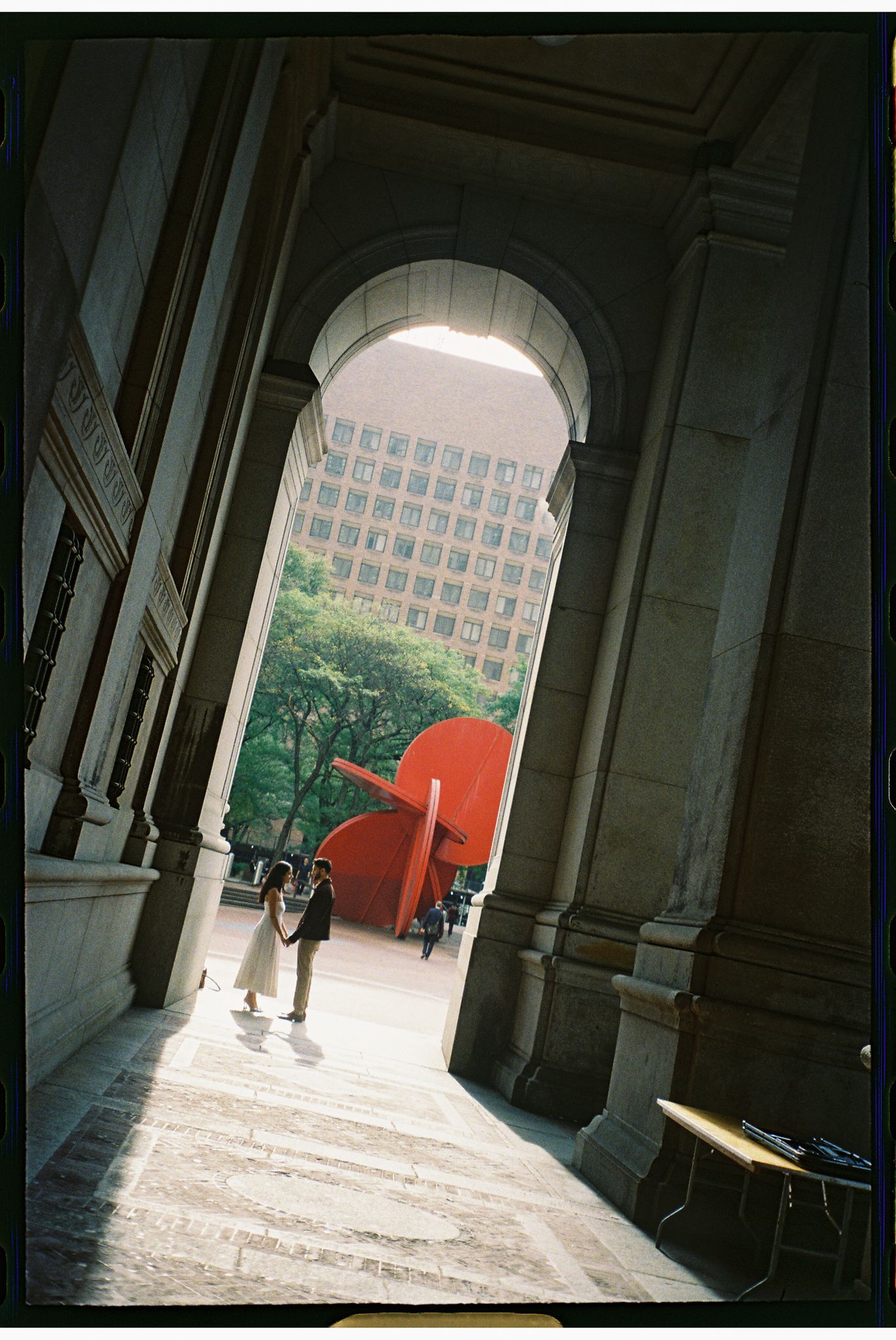 A series of architectural columns frame a walkway leading to a bright outdoor space with a red sculpture visible in the distance.