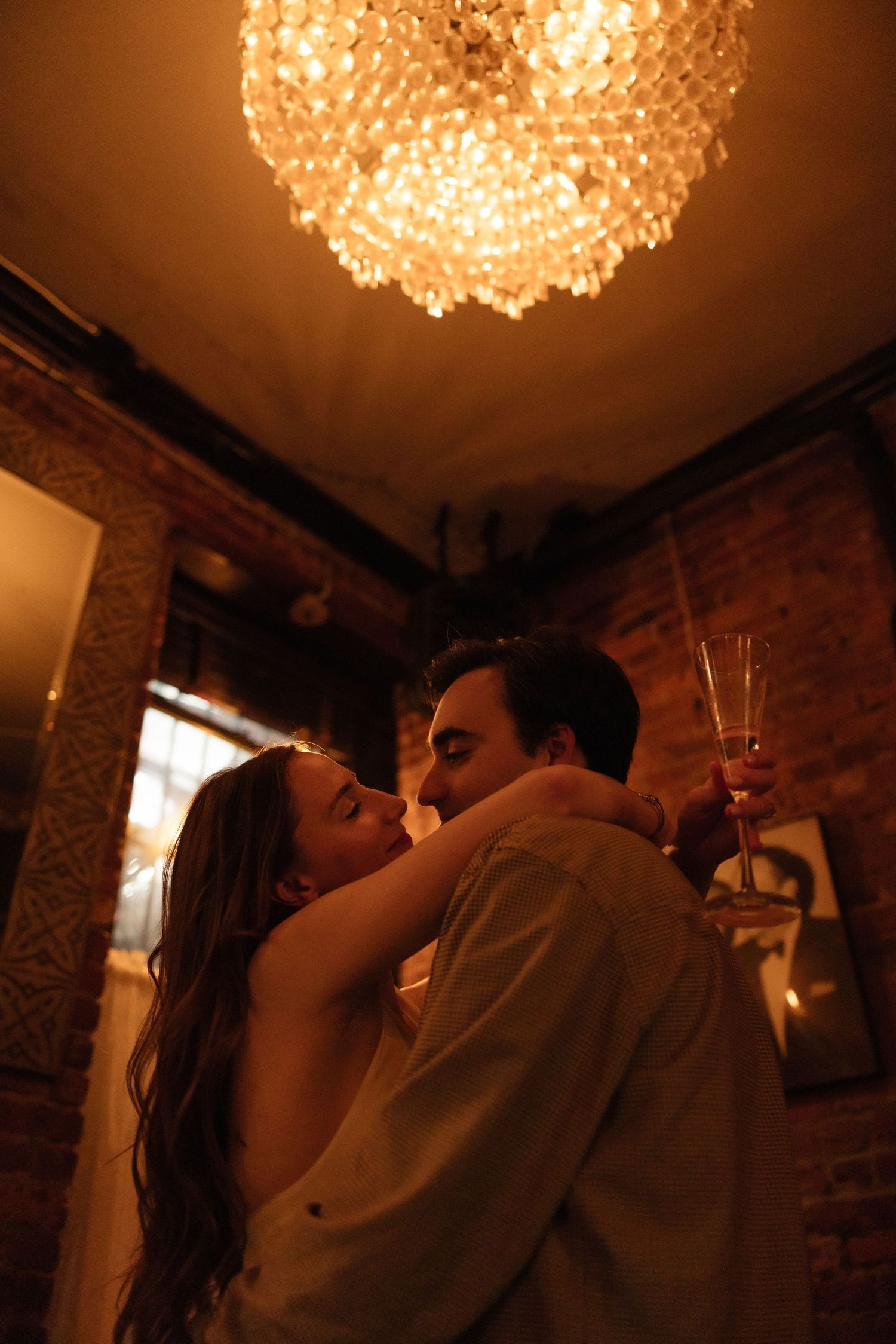 A romantic couple shares an intimate dance moment under a warm crystal chandelier in a rustic brick-walled venue.