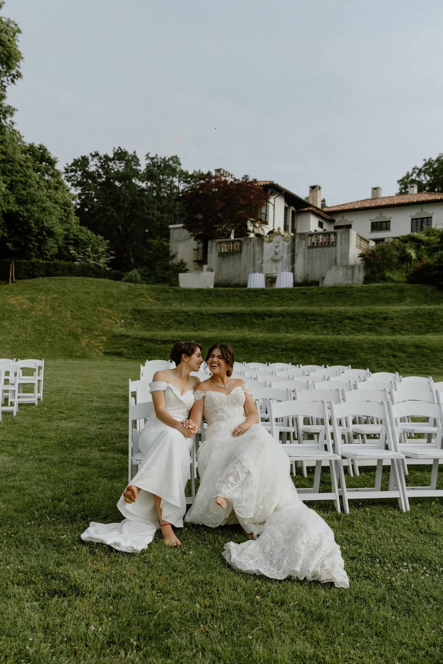 Two brides in white gowns sit together on a lawn at an outdoor wedding ceremony with white chairs and an elegant estate.