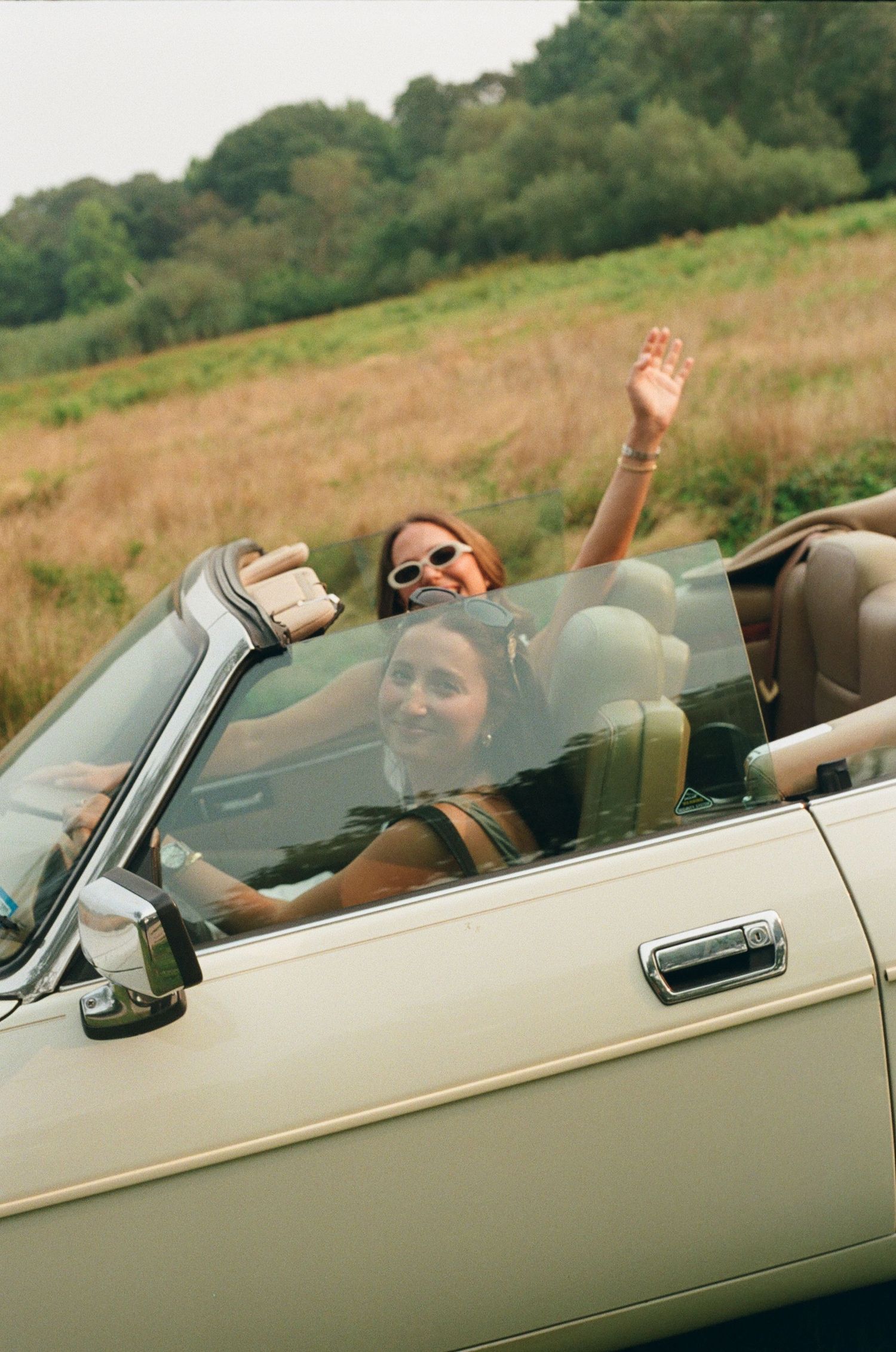 People enjoying a summer drive in a vintage convertible car through a scenic countryside with rolling hills in the background.