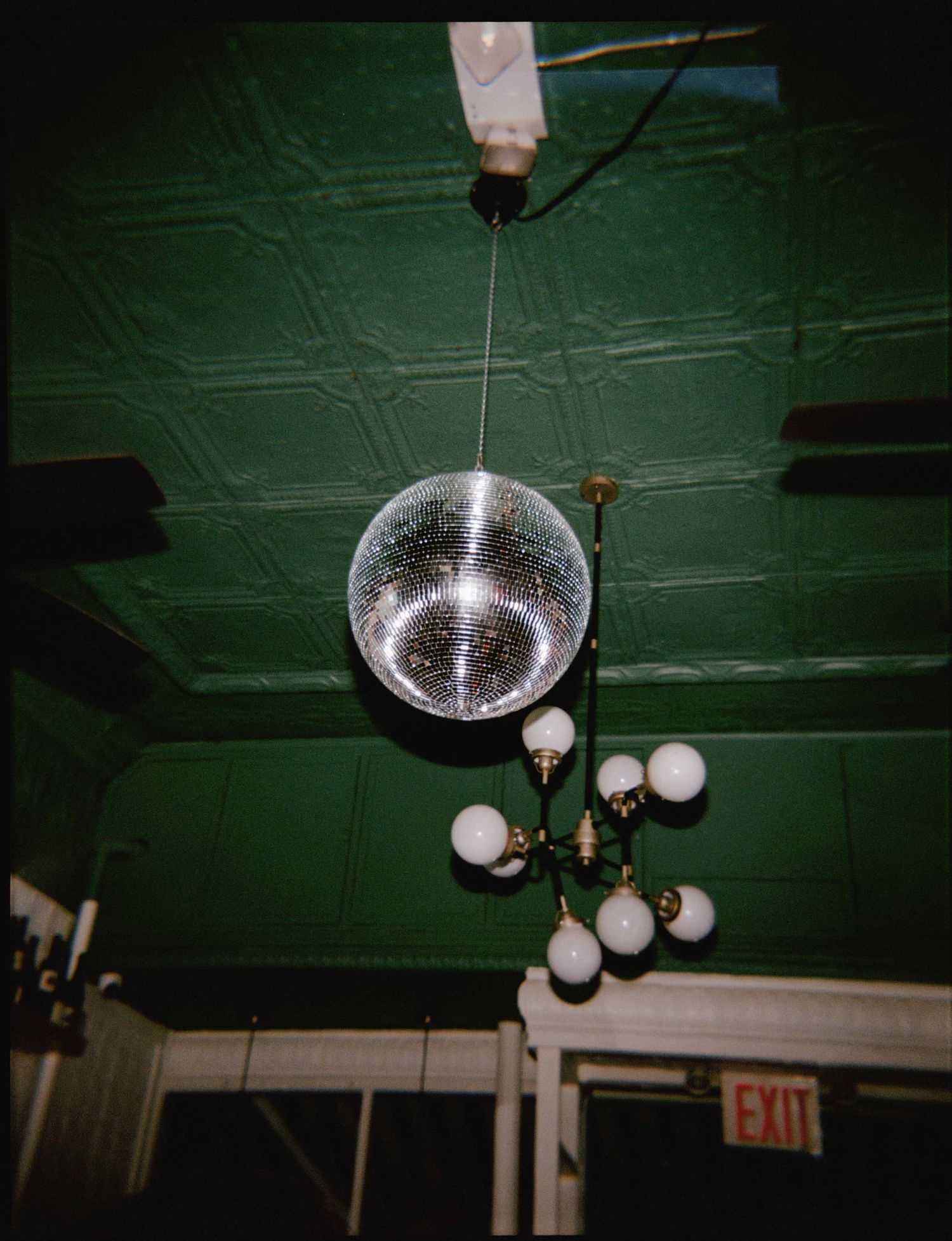 A silver disco mirror ball hangs from a green pressed-tin ceiling beside a vintage globe chandelier, with an exit sign glowing below.