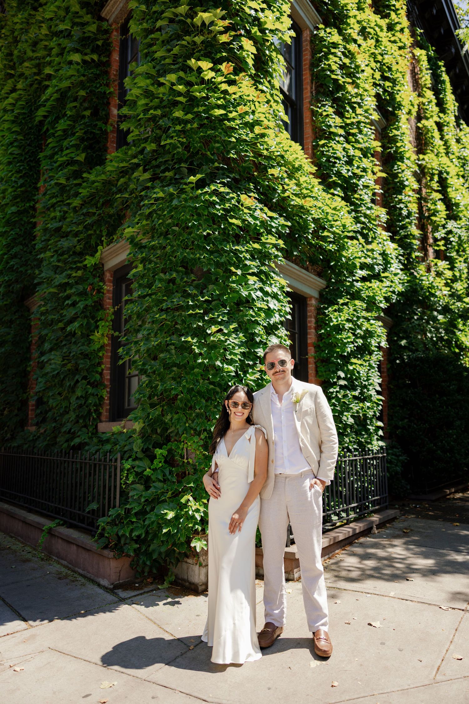 Bride and groom in white and beige attire pose on a sunny sidewalk in front of an ivy-covered brick building.