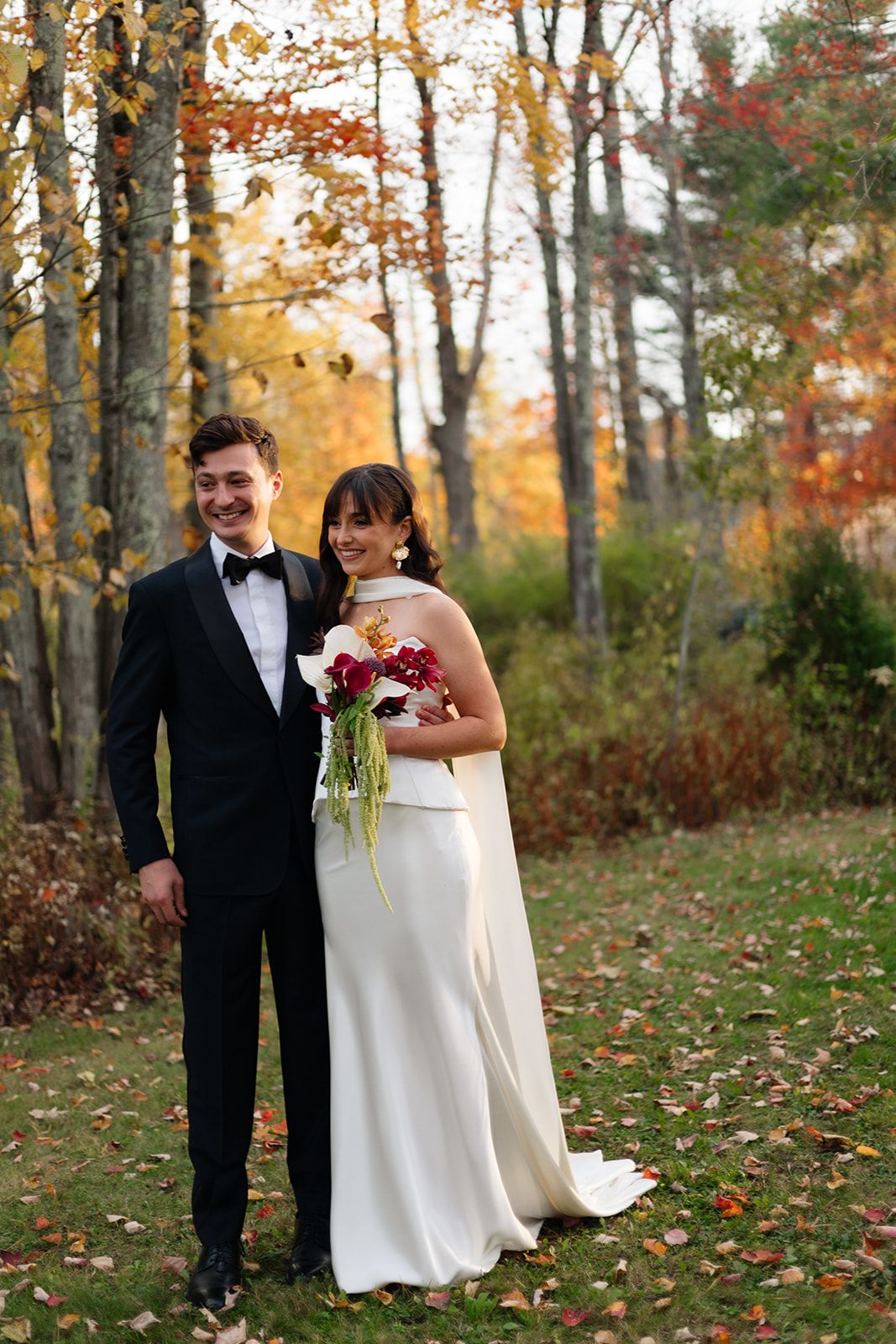 A couple in wedding attire poses in a wooded area with autumn foliage and fallen leaves on the ground.