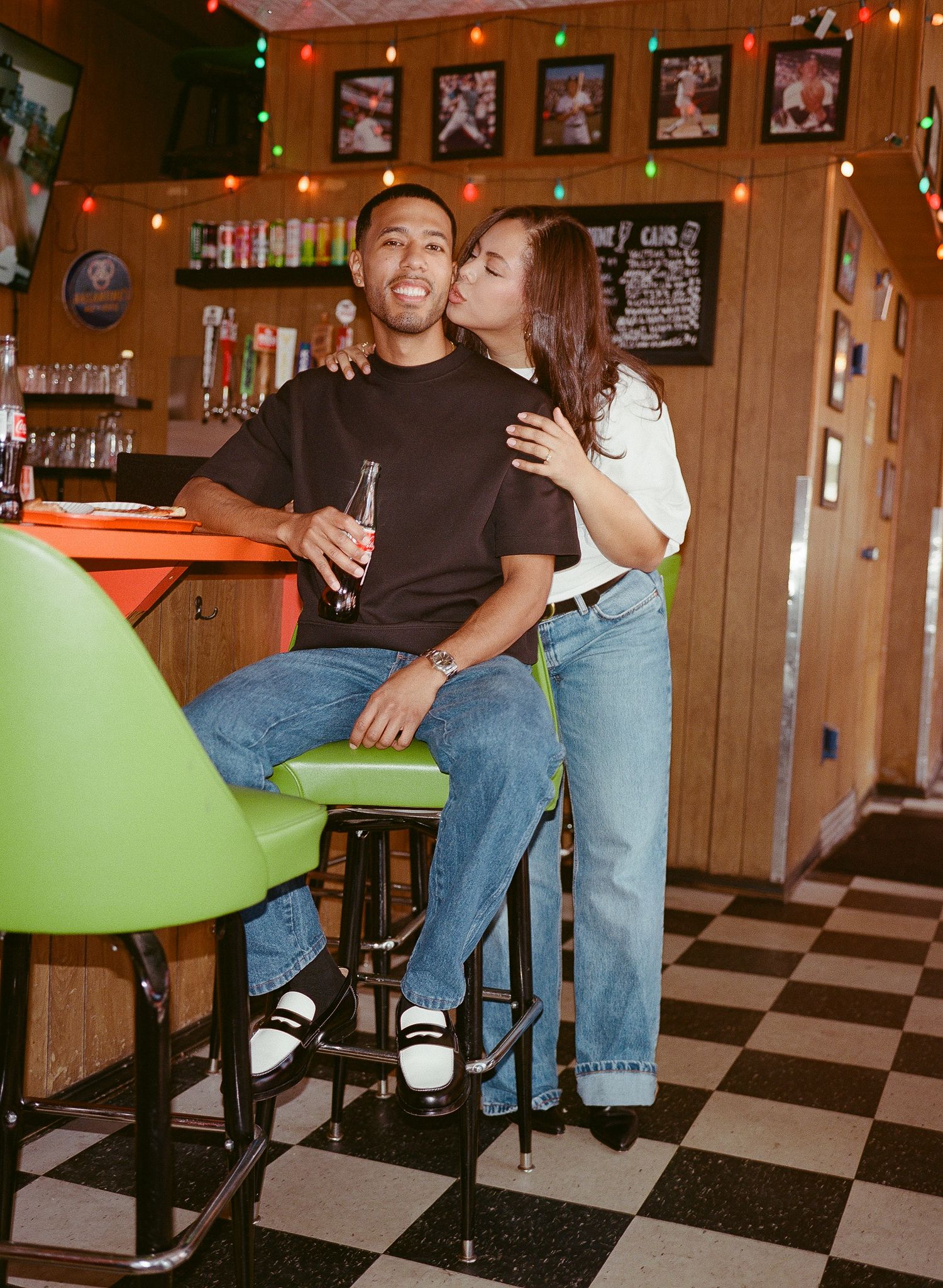 People sitting at a retro diner counter with black and white checkered floor and green booth seats in warm lighting.