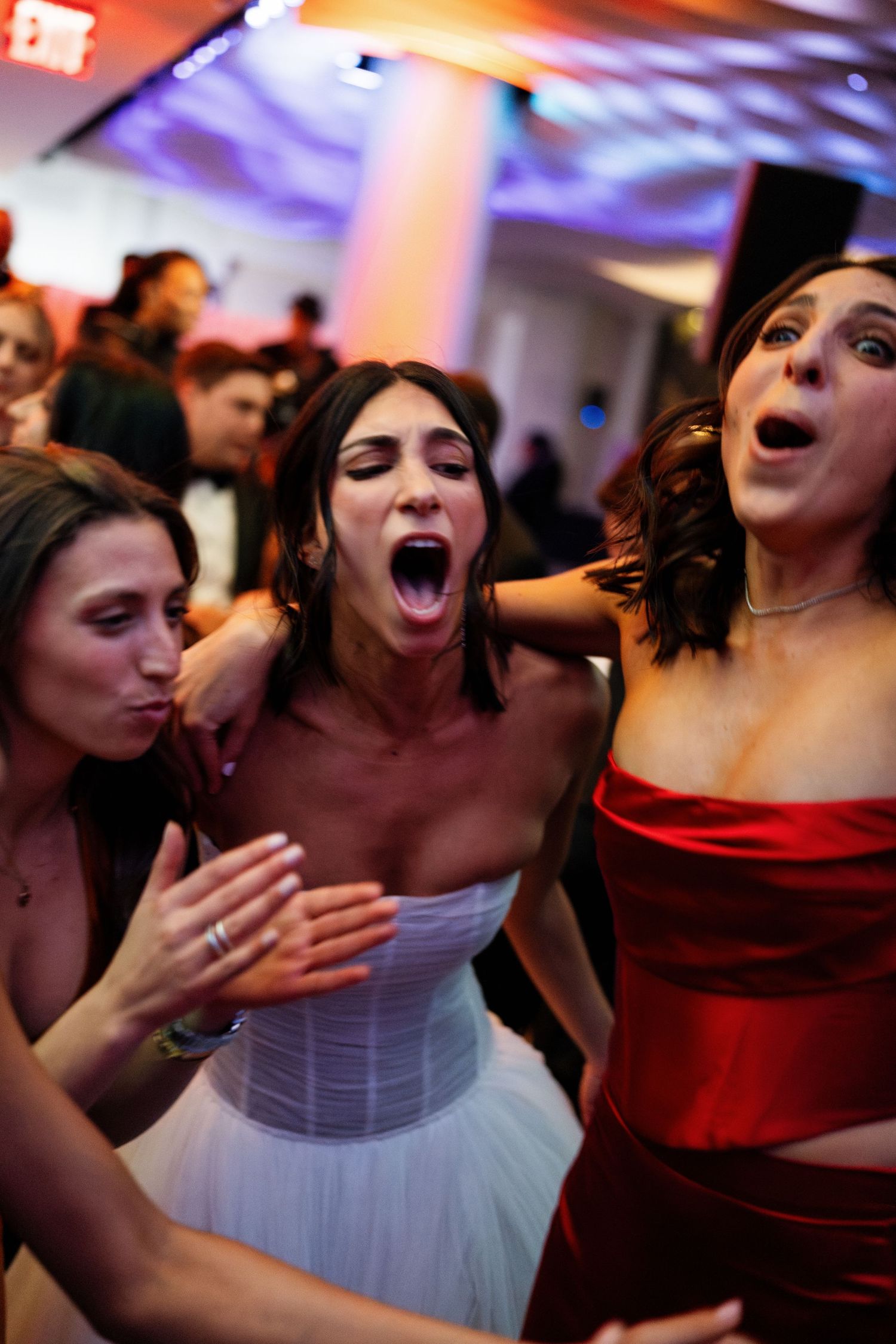 Bride in white corset dress dancing joyfully with friends in red dress at lively wedding reception.