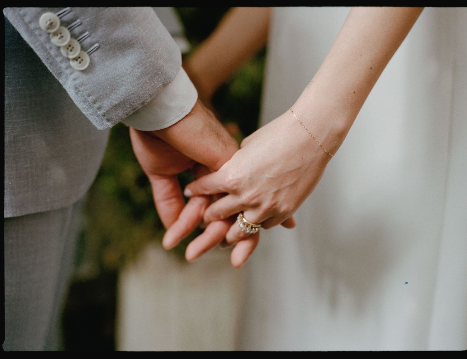 Close-up of a couple holding hands, the bride wearing a white dress and a diamond ring, the groom in a gray suit with button cuffs.