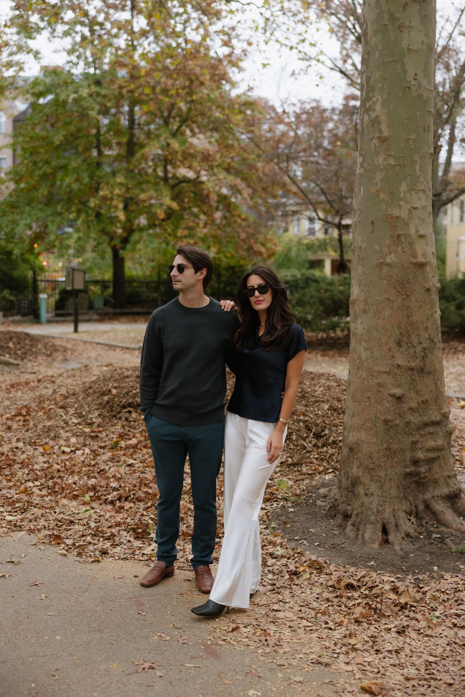 A couple sits together on fallen autumn leaves in a park with tall trees on a cool fall day.