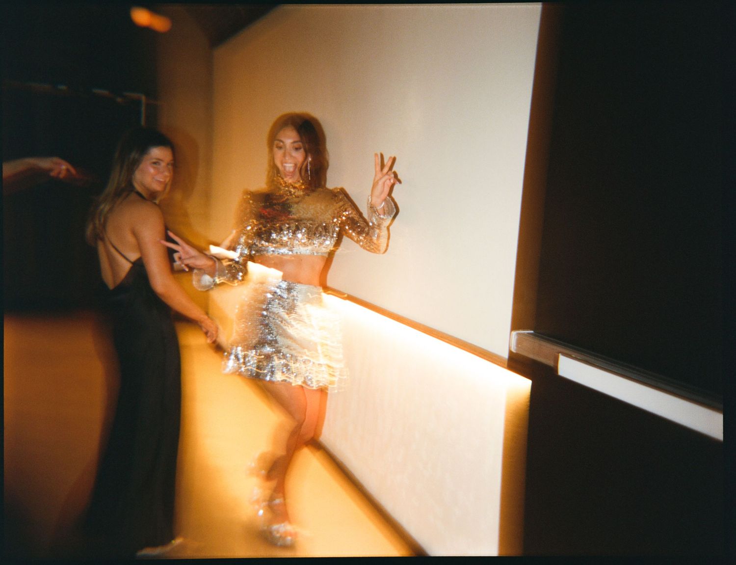 Two women pose in a dimly lit venue, one wearing a sparkling silver mini dress and the other in a black backless gown.