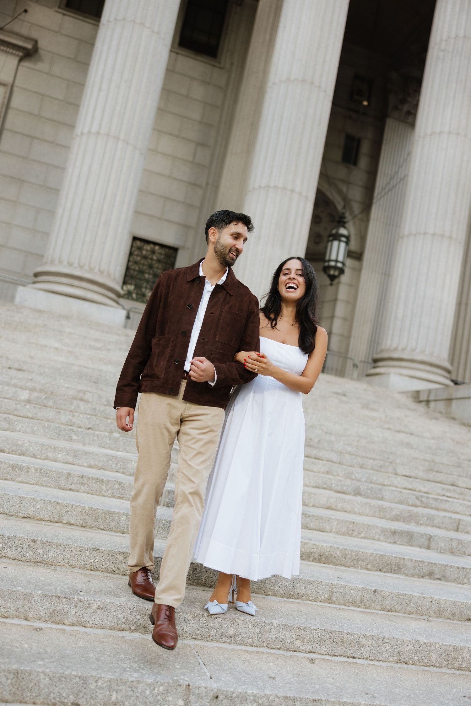 A couple laughs together while descending white marble steps of a building with classical columns.